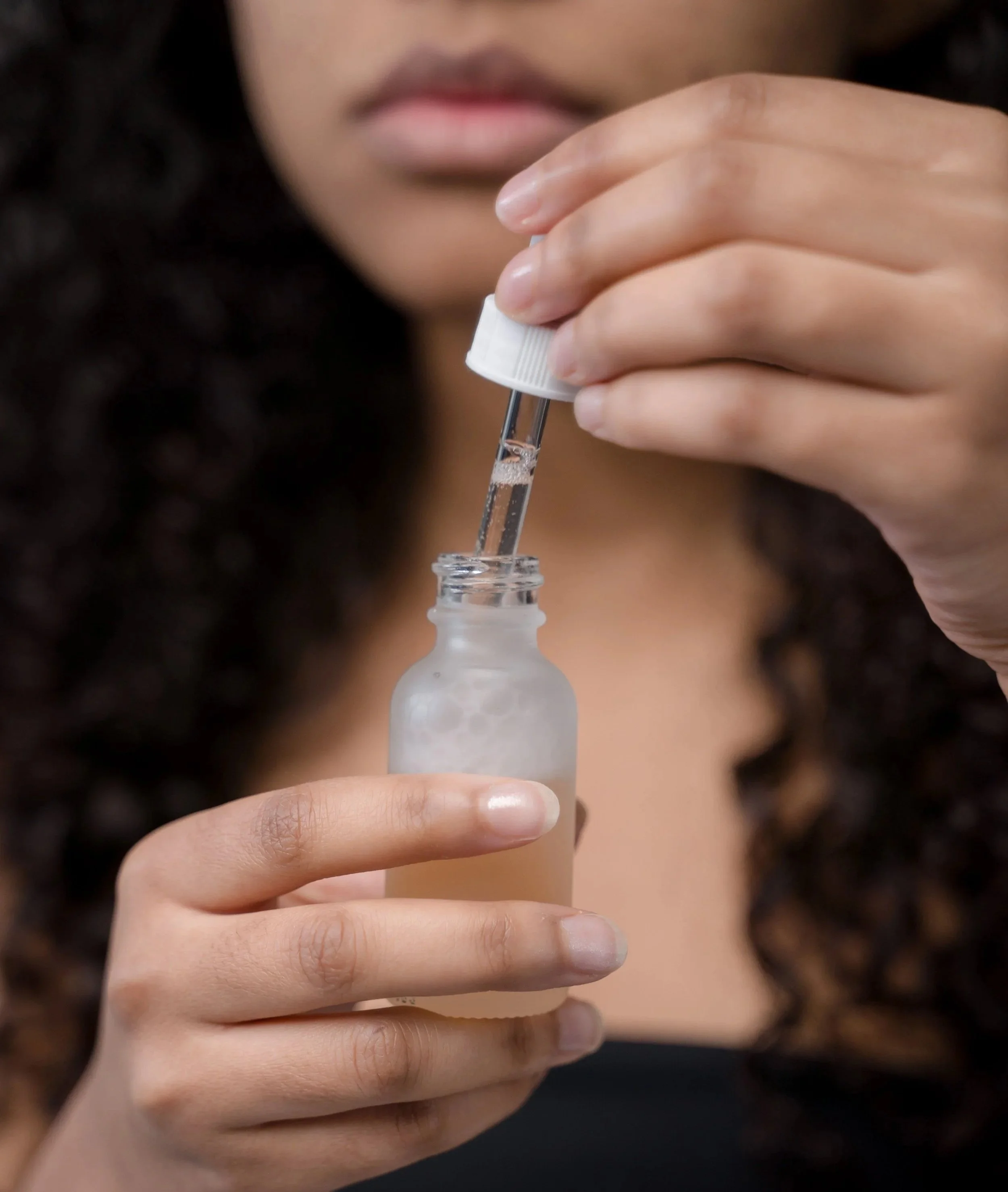 Close-up of a woman with curly dark hair holding a glass dropper and serum bottle, illustrating the use of scalp serums and hair care treatments for healthier-looking hair, with hair care insights from TEAL Hair & Makeup Studio.