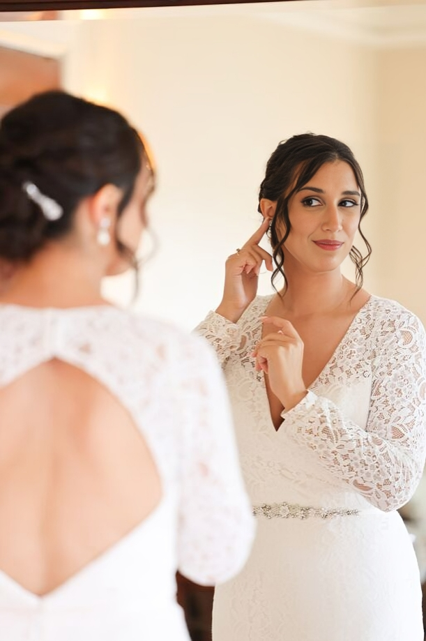 Bride in a lace wedding gown viewing her finished bridal hair and makeup in a mirror before the ceremony.