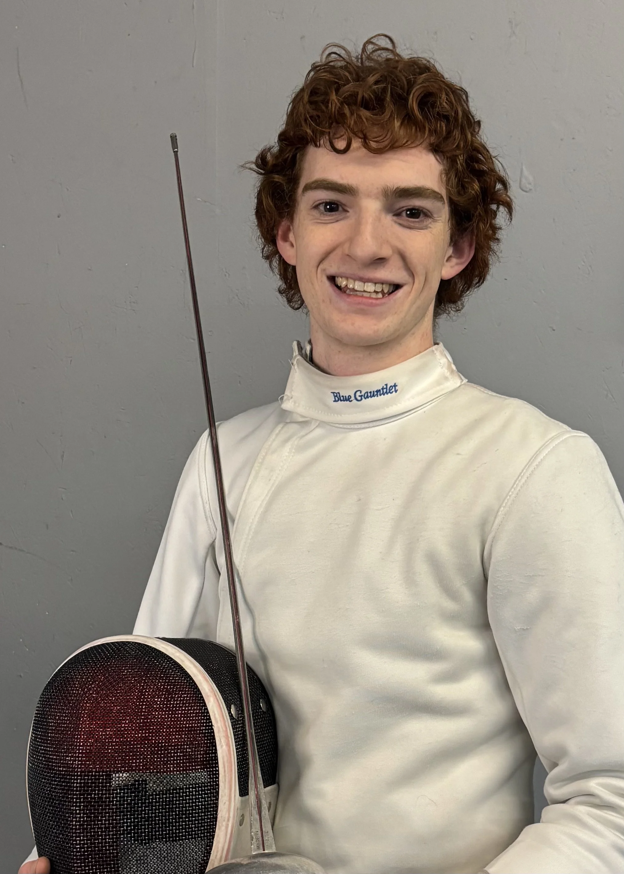 Young man with curly red hair smiling in fencing gear, holding a fencing mask and foil, standing against a gray wall.