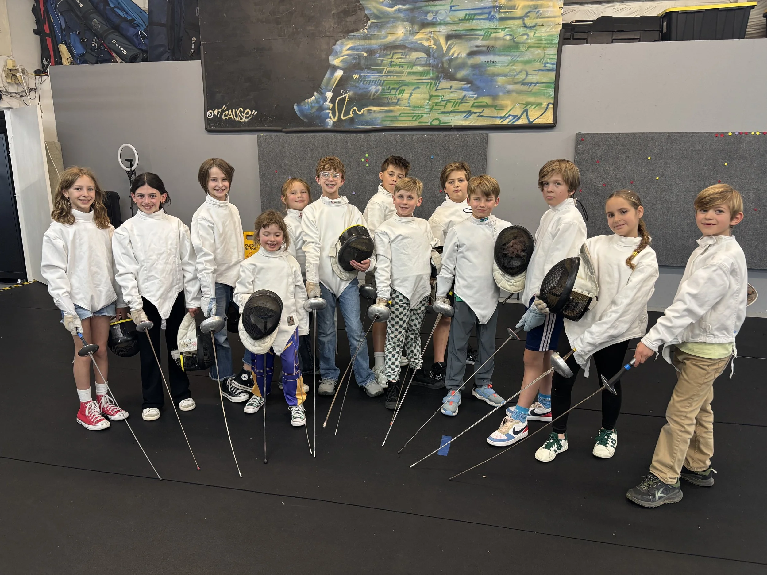 Group of children dressed in fencing gear, holding fencing helmets and swords, standing in an indoor fencing gym.Northern Colorado Fencers - Fencing, fencing for children