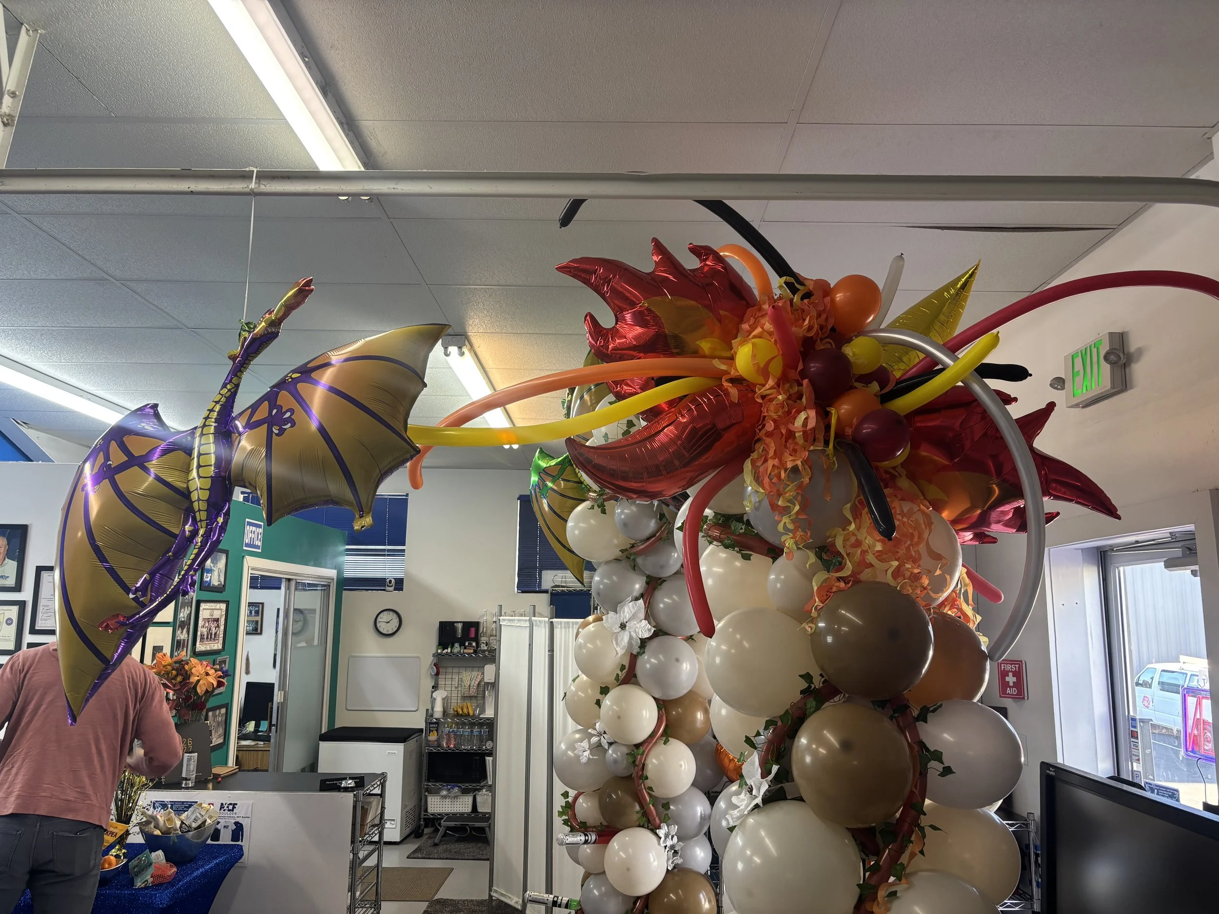 Colorful balloon arrangement with gold and purple dragon-shaped balloon and a multicolored balloon cluster with red, orange, yellow, white, and brown balloons inside an office.