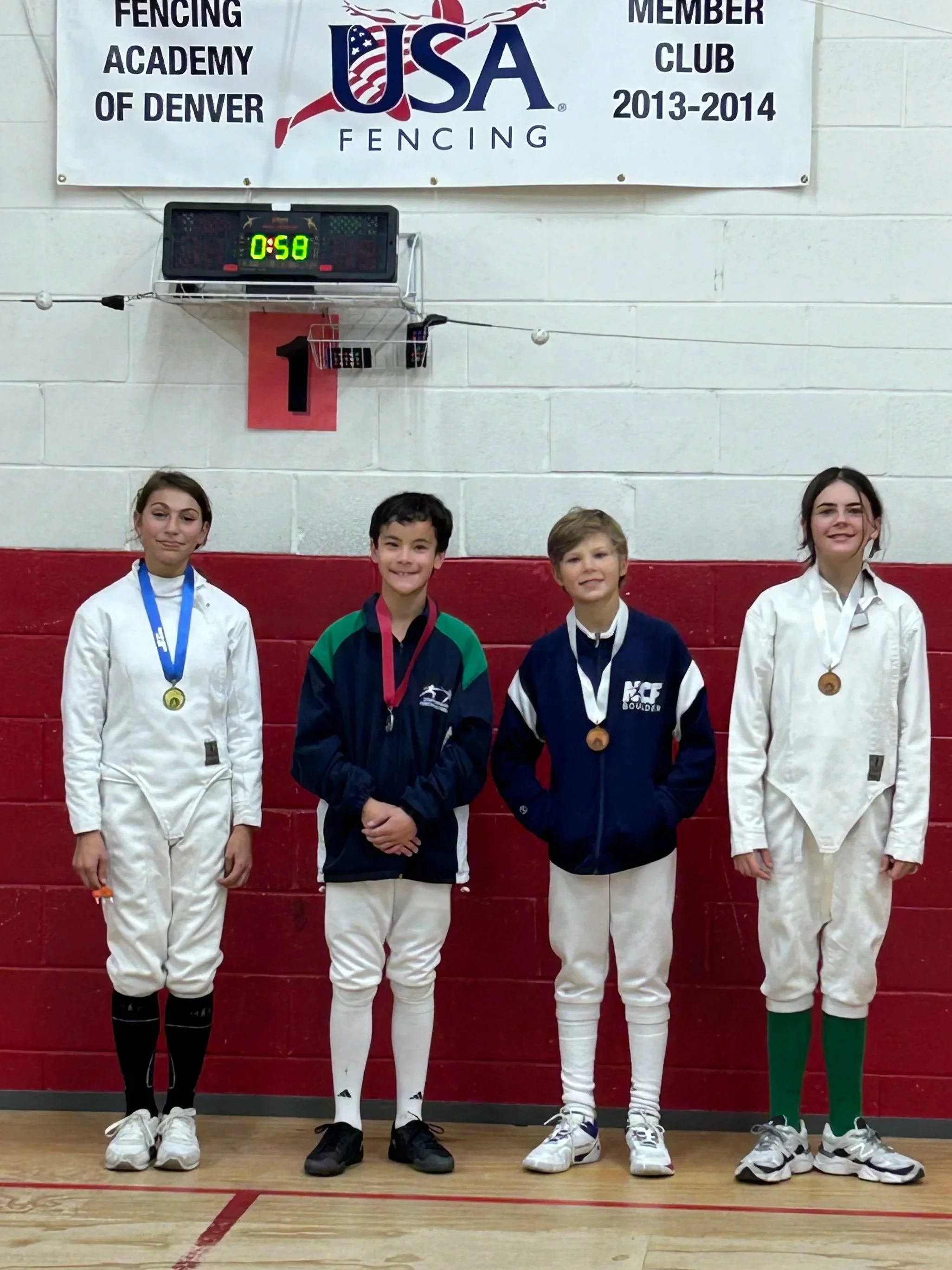 Four young athletes standing in a gymnasium in fencing uniforms and medals, with a scoreboard and banner in the background, celebrating a fencing competition. The banner reads 'USA Fencing, Fencing Academy of Denver, Member Club, 2013-2014'.