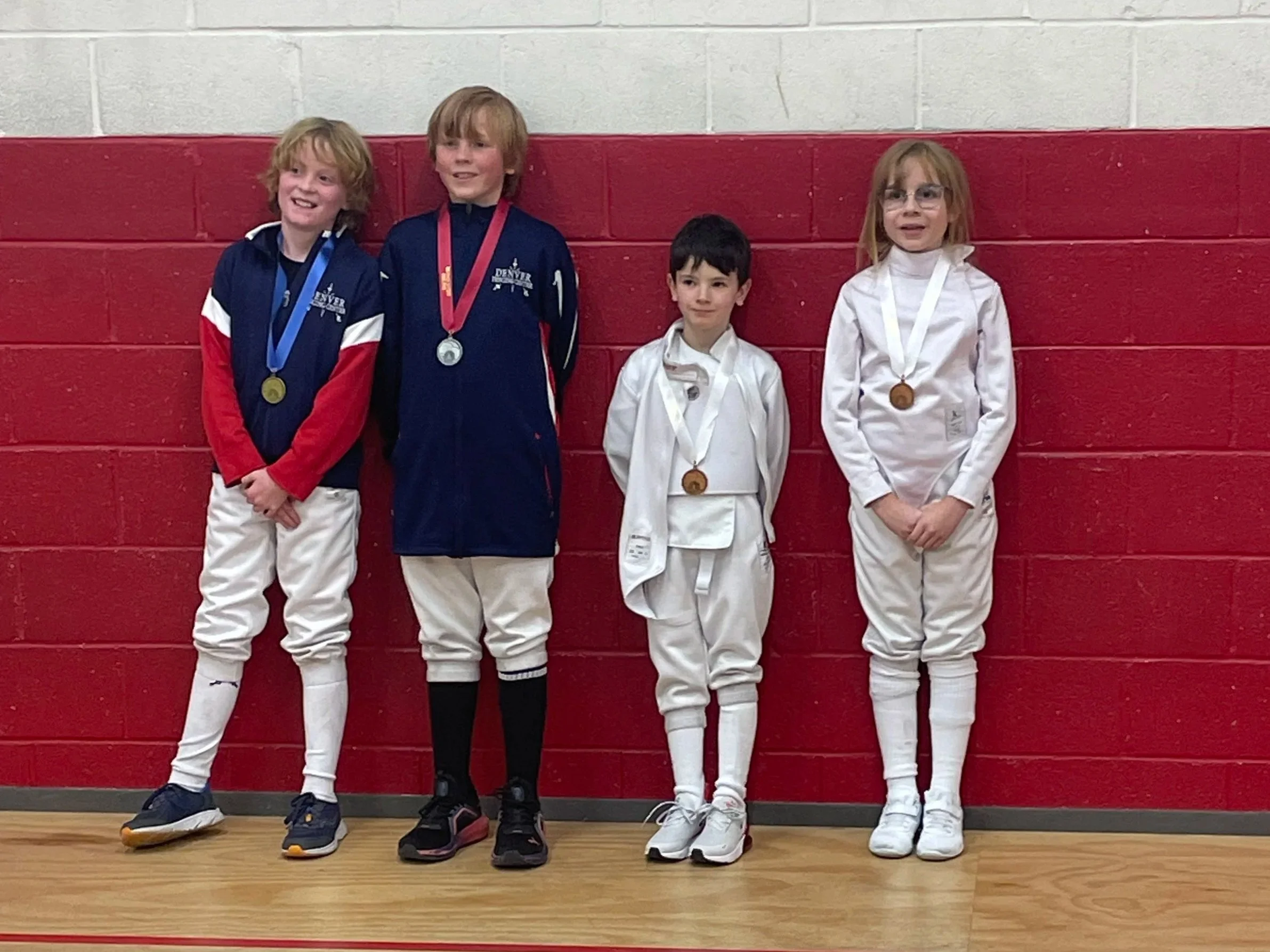 Four young children standing against a red and white wall, wearing fencing uniforms and medals around their necks.