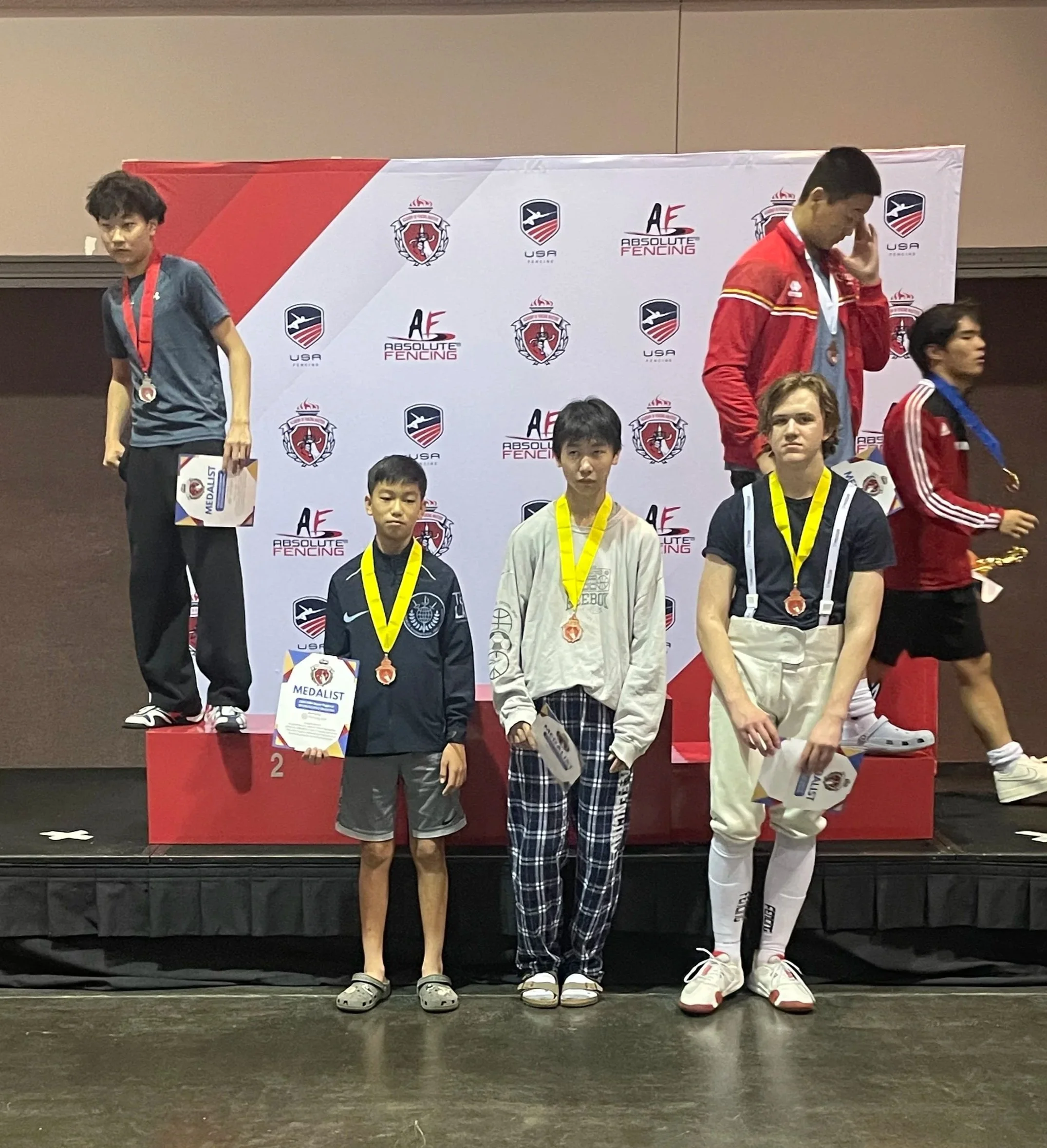 Kids standing on a winners' podium at a fencing competition, holding medals and certificates, with a backdrop featuring fencing organization logos.