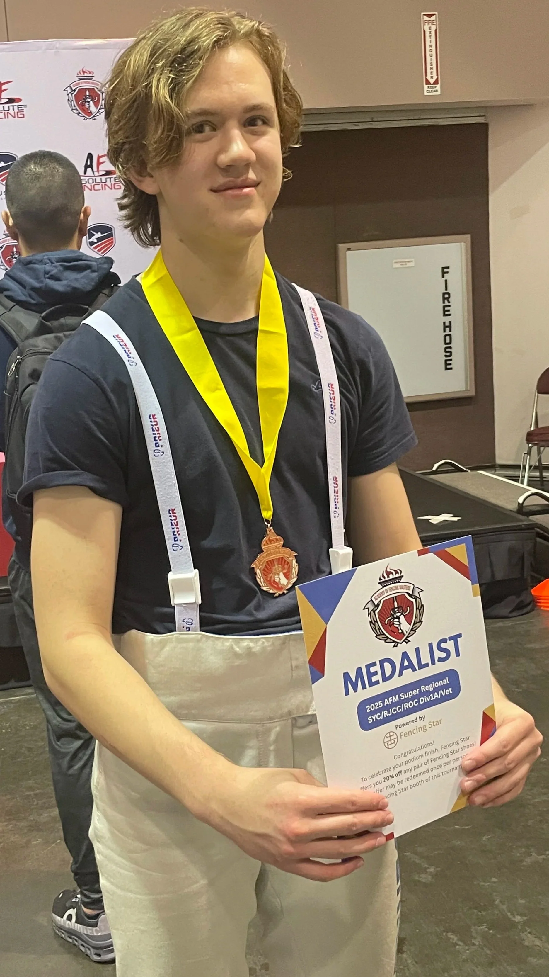 Young male athlete with curly hair wearing a black t-shirt, beige fencing jacket, and a gold medal around his neck, holding a certificate that reads 'Medalist' at a fencing competition awards ceremony.