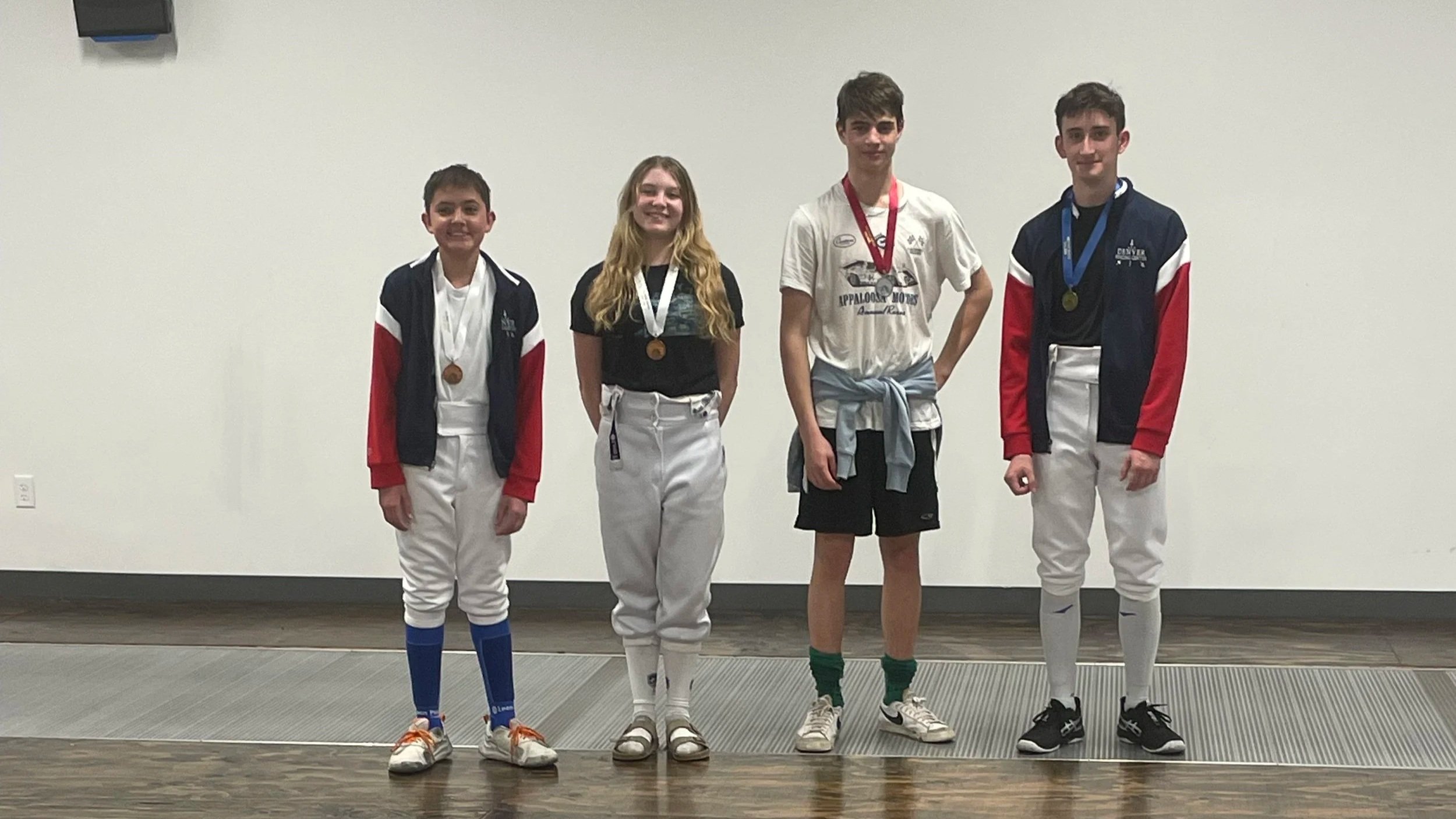 Four young athletes standing on a podium, each wearing medals, with a plain wall background and wooden and metallic flooring.