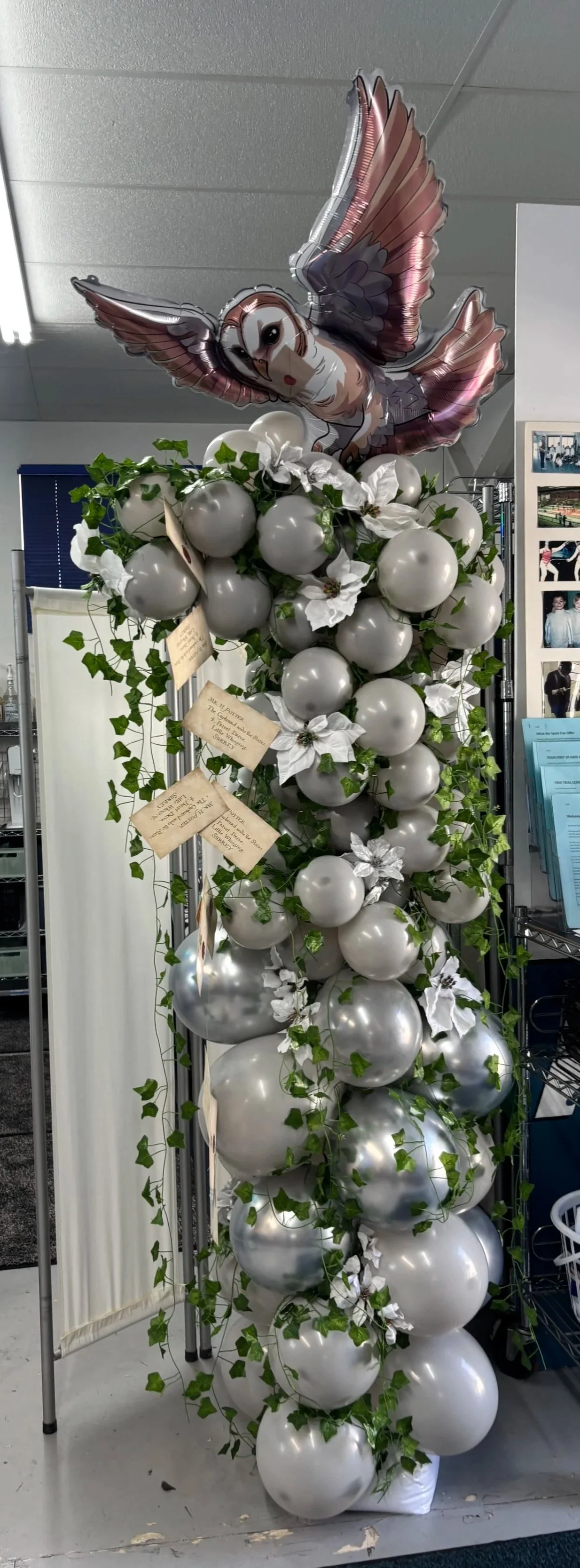 A decorative arrangement featuring a large bouquet of white balloons intertwined with green ivy and white flowers, topped with a balloon of an owl with outstretched wings.