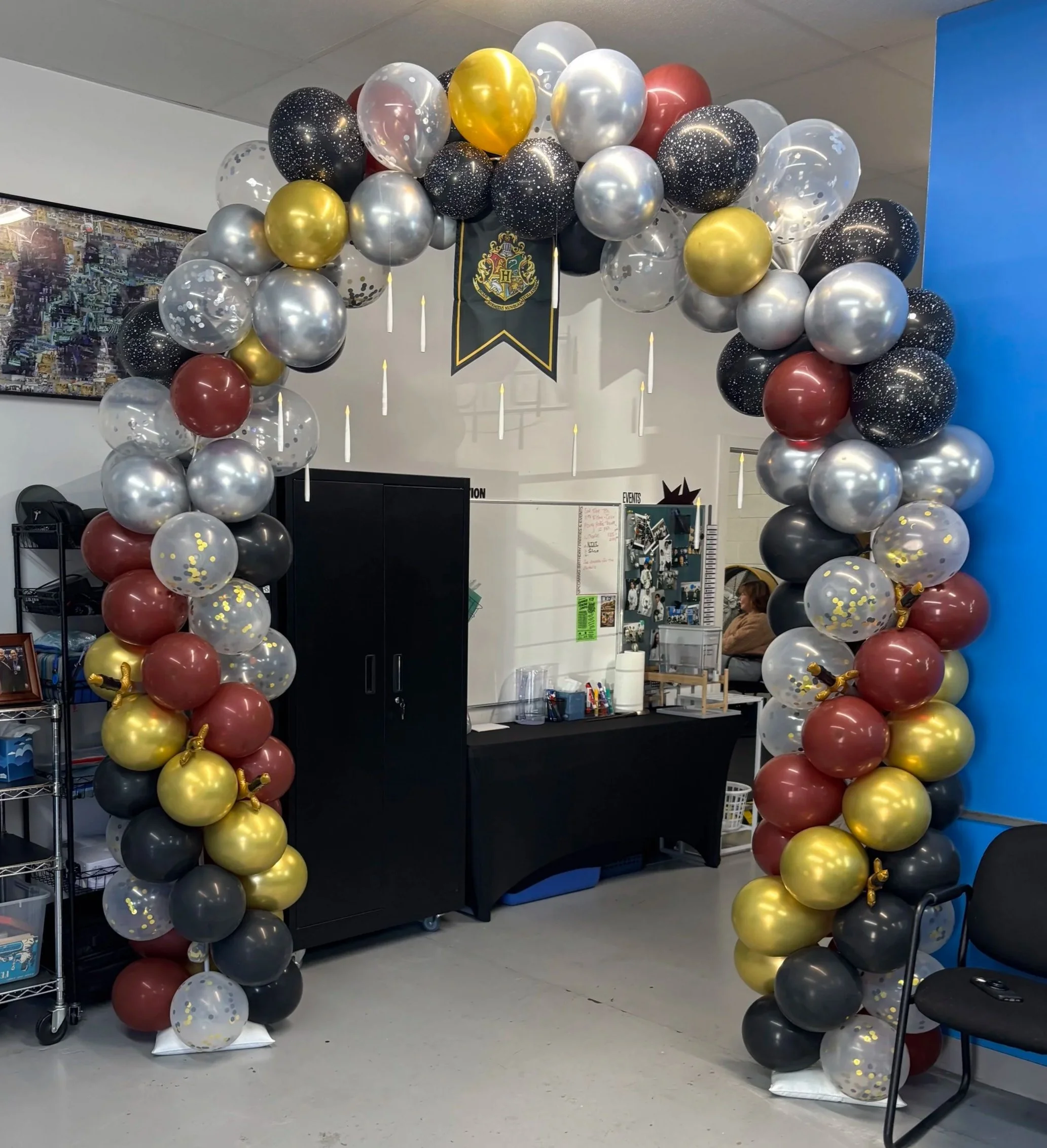 Balloon arch with black, gold, red, and transparent balloons decorated with gold confetti, featuring a Hogwarts crest banner hanging in an indoor room, likely for a celebration or event. Boulder birthday party.