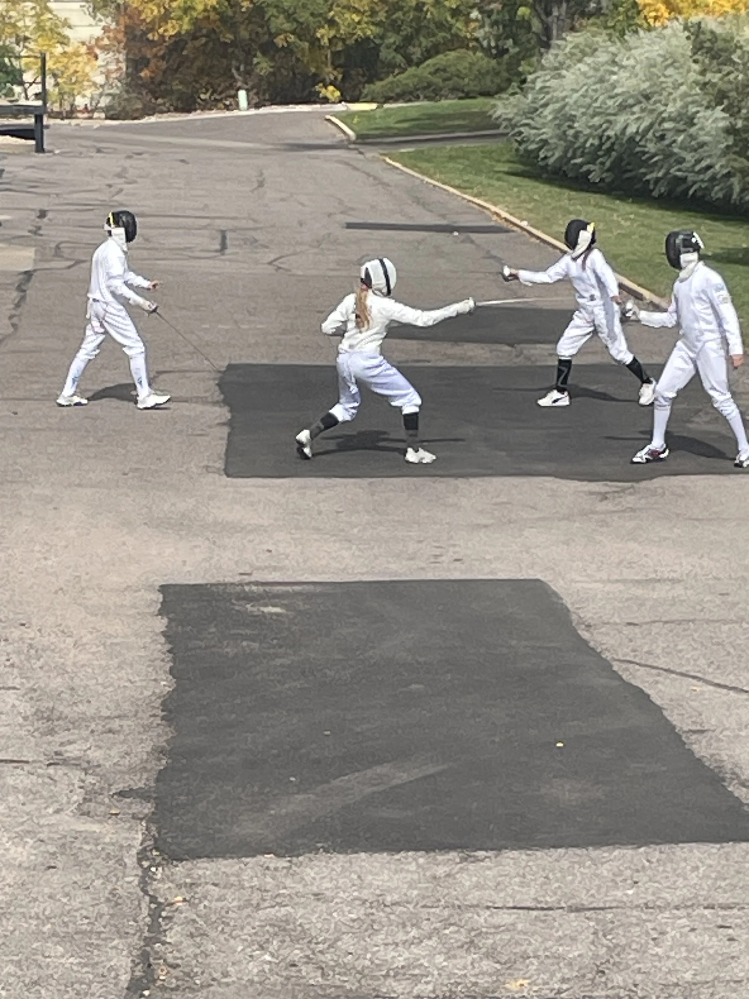 Group of four people dressed in fencing gear practicing fencing outdoors on a paved area with black patches, with trees and greenery in the background.