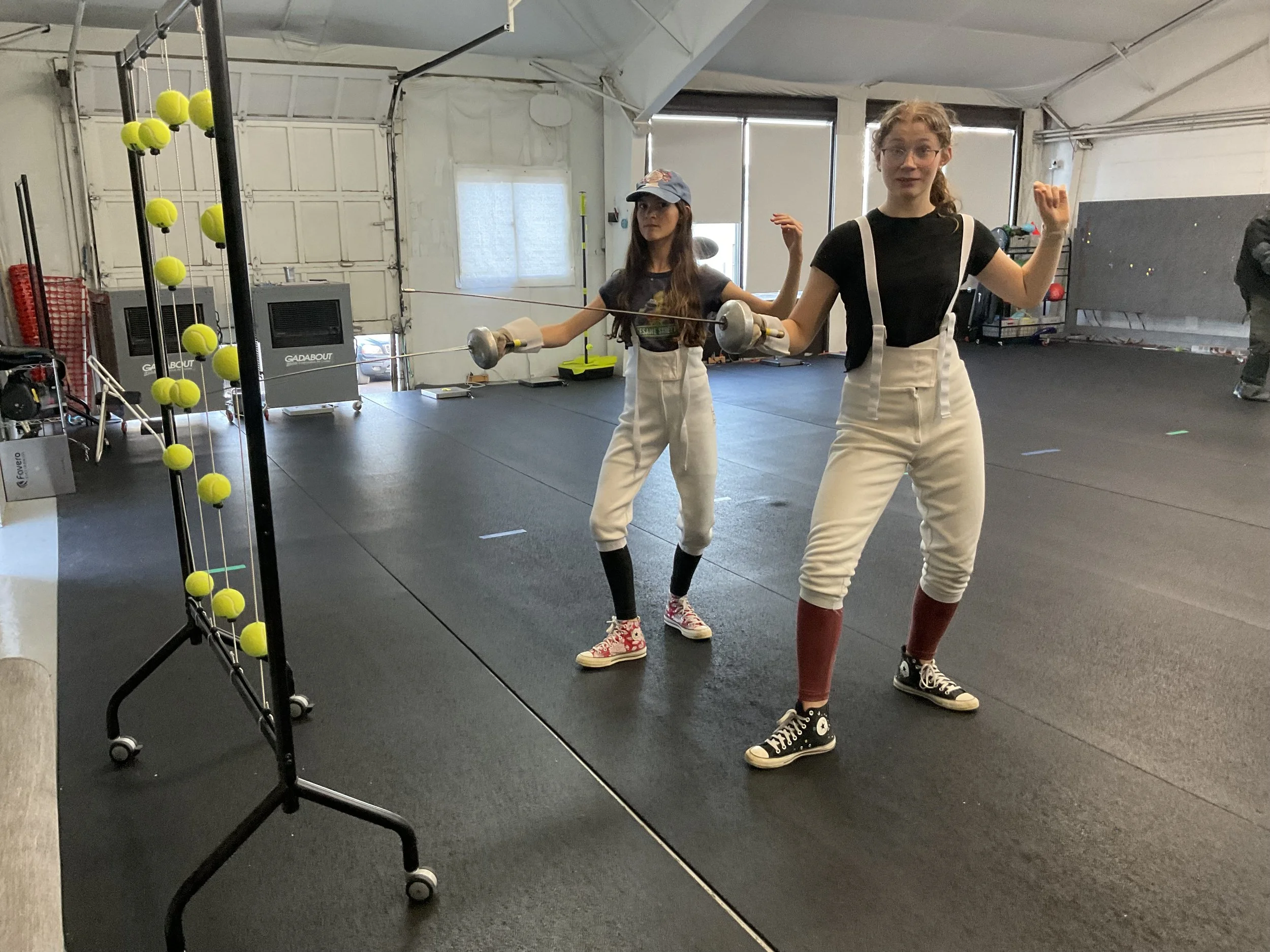 Two young women practicing fencing indoors in a gym, wearing vintage-style baseball jerseys, high-waisted white pants, and sneakers, with one in front holding a fencing foil and both posing dynamically.