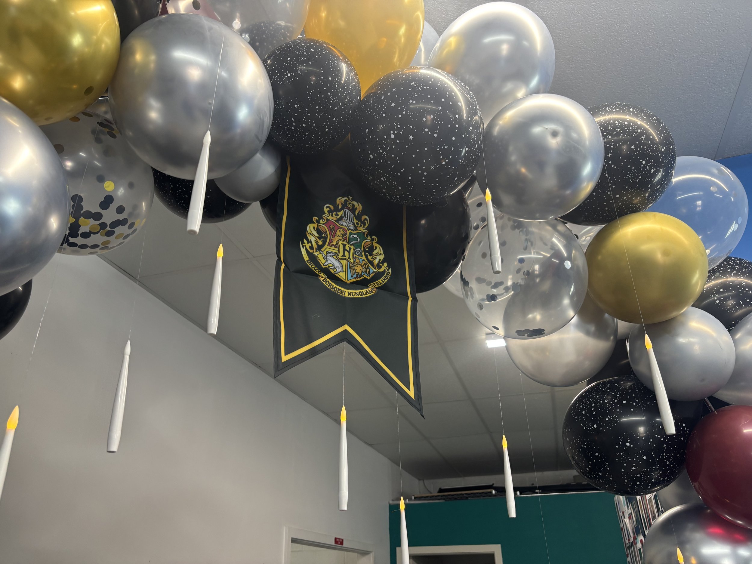 A large balloon display with black, gold, silver, and confetti-patterned balloons hanging from the ceiling, featuring a Hogwarts crest banner in the center, and candle-shaped balloons interspersed among them.