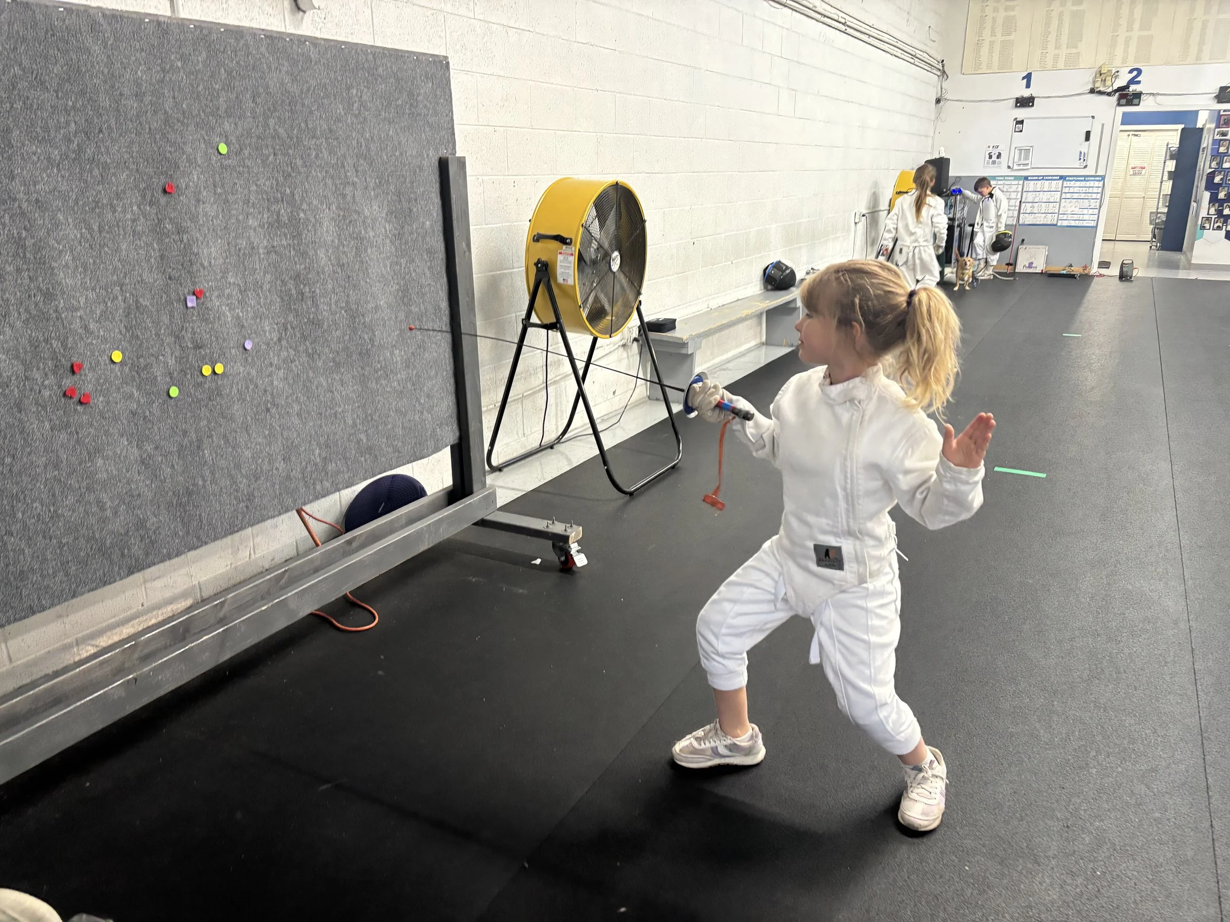 A young girl dressed in a white fencing uniform practicing fencing with a foiled sword in a martial arts or fencing gym.
