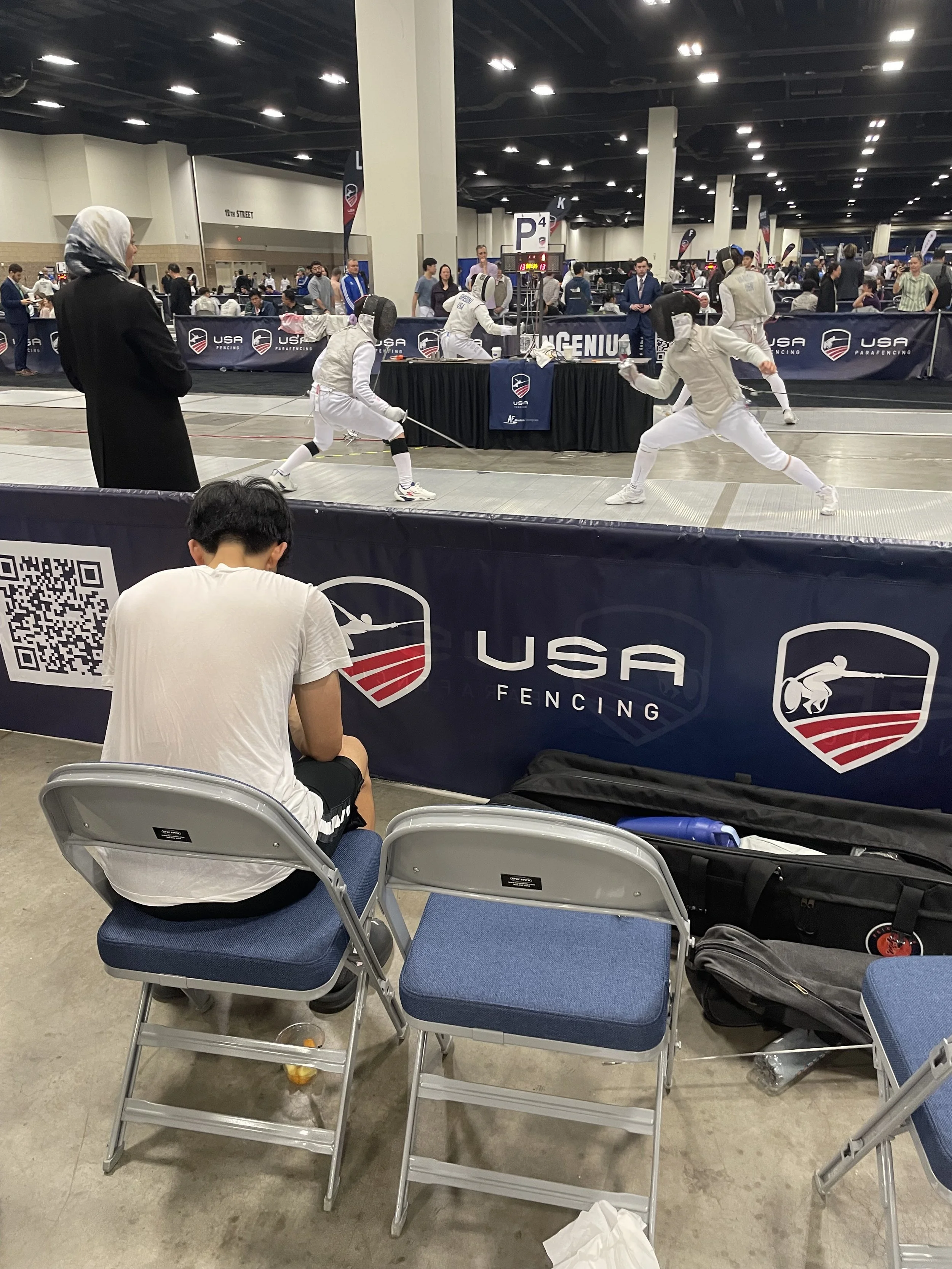 A youth fencing match taking place at a USA Fencing competition in an indoor arena, with two fencers in white protective gear fighting, a referee observing, and spectators watching from behind a barrier.