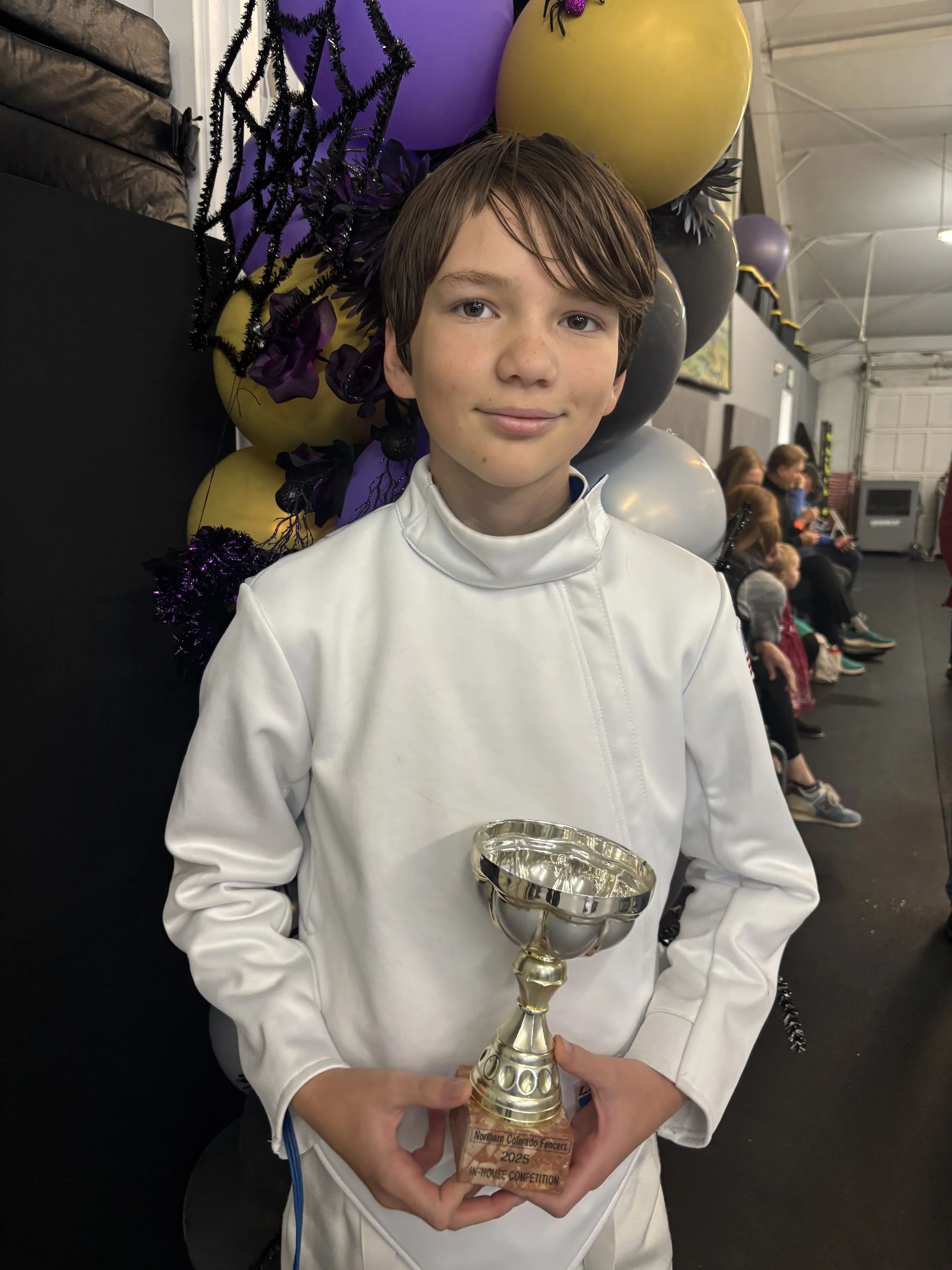 A young boy with short brown hair holding a silver trophy with a wooden base. He is wearing a white fencing uniform and standing in front of a colorful decoration with purple, yellow, black, and gray balloons and black tinsel.