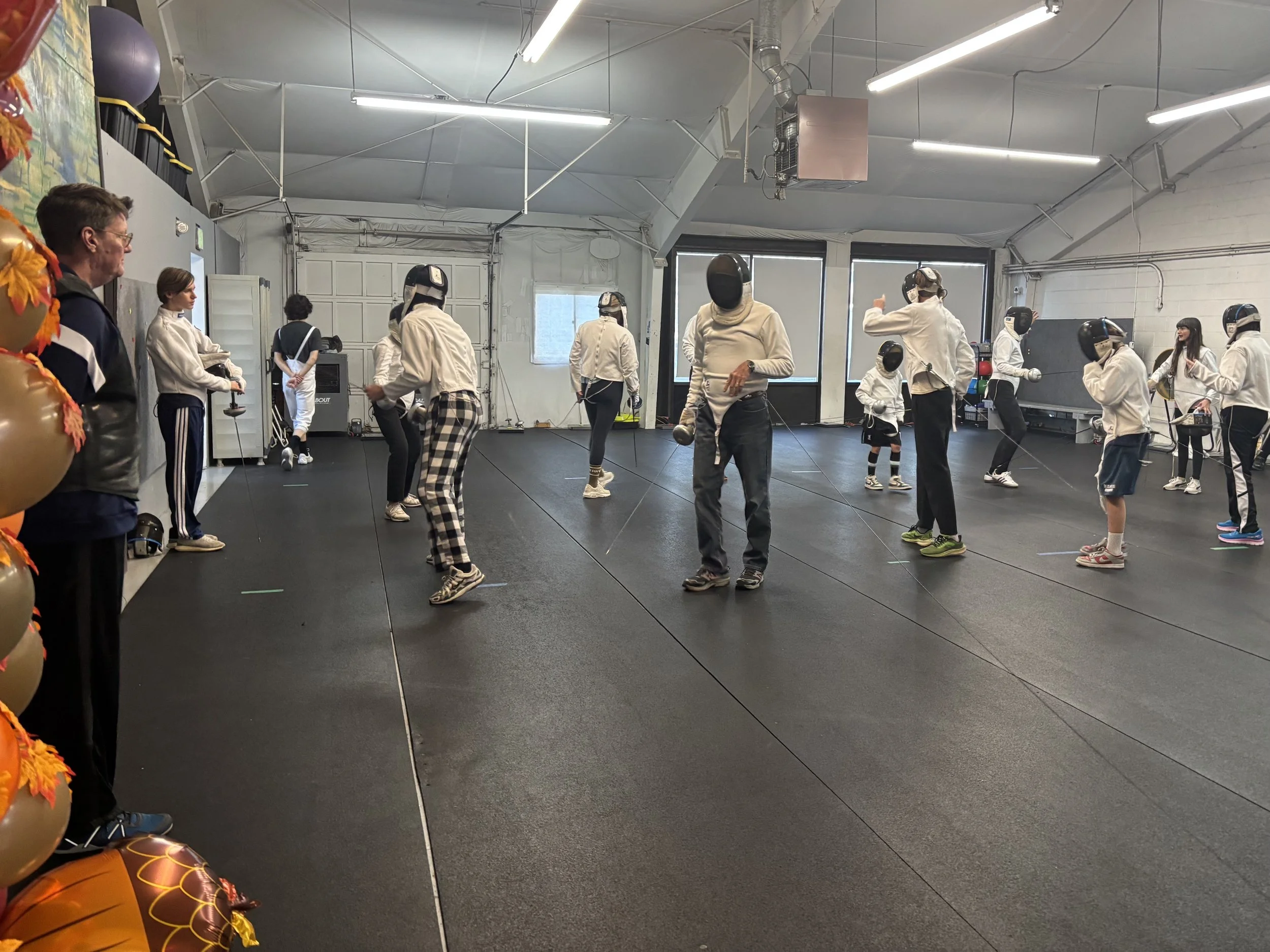 People in white fencing masks and protective gear practicing fencing in an indoor gym.Youth fencing in Boulder at Northern Colorado Fencers.