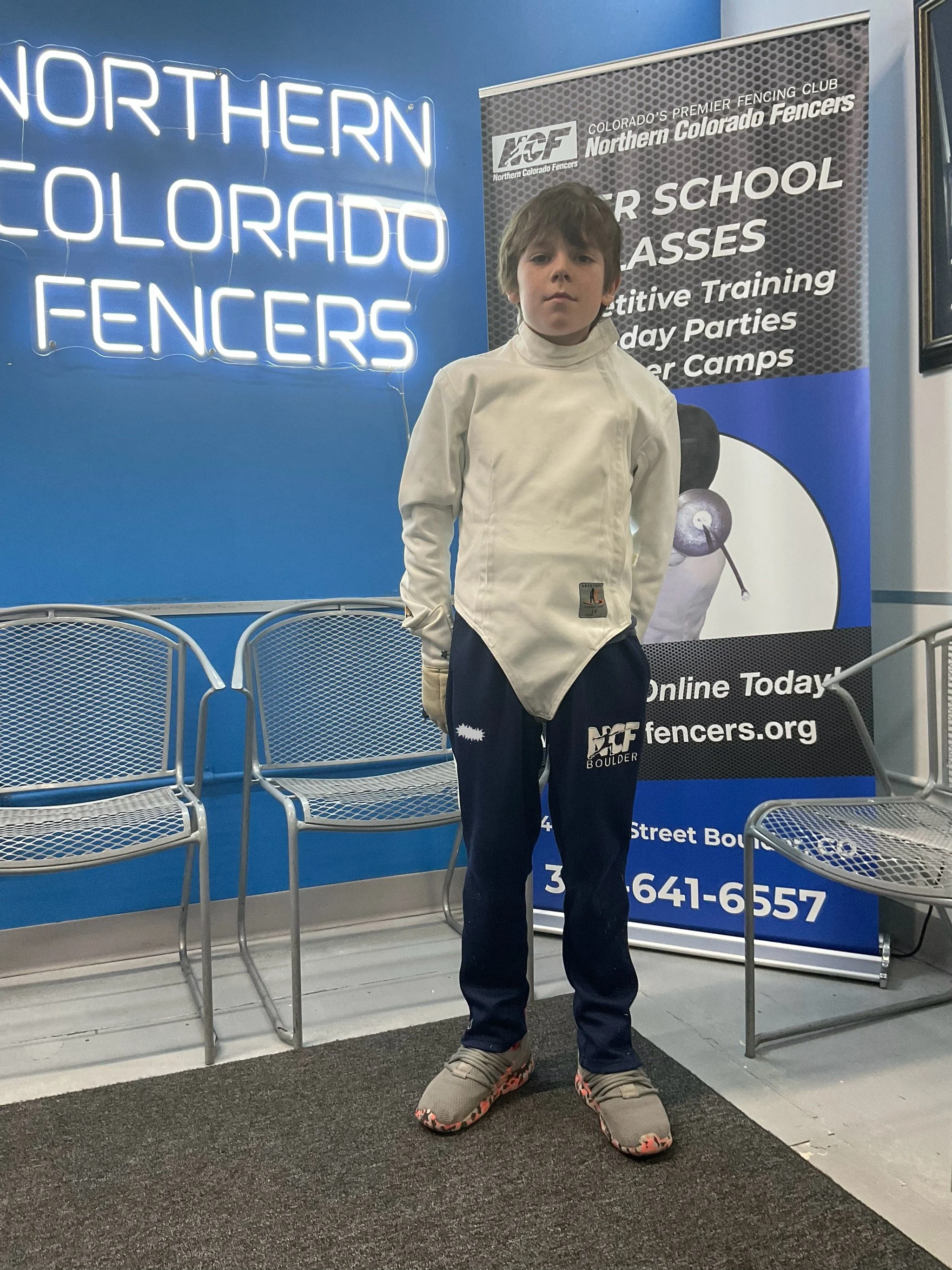 Young boy in fencing outfit standing in front of a Northern Colorado Fencers sign and banners at a fencing club.