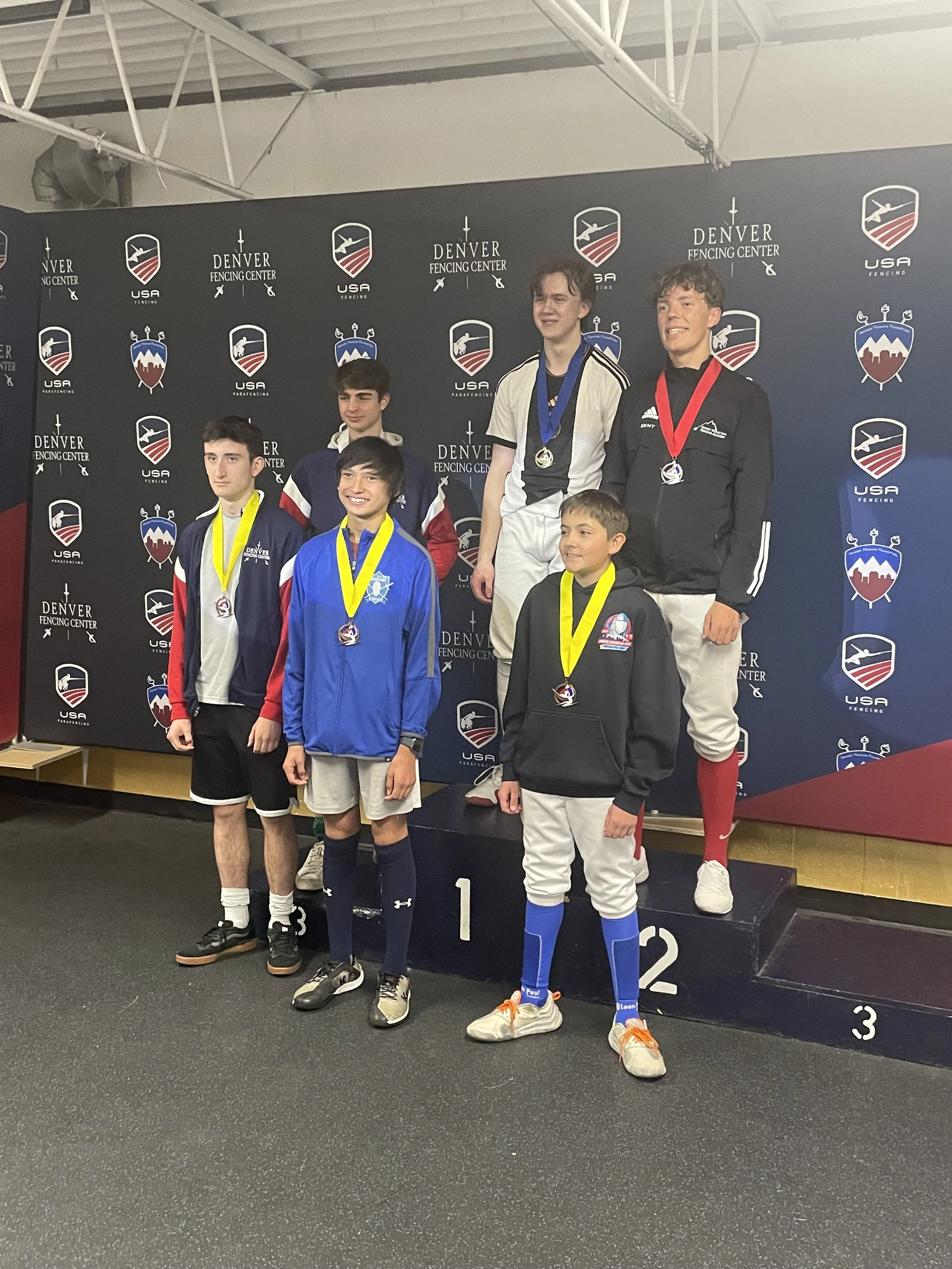 Young male athletes standing on a winners' podium with medals at a fencing competition, with a backdrop displaying 'Denver Fencing Center' and 'USA Fencing' logos.