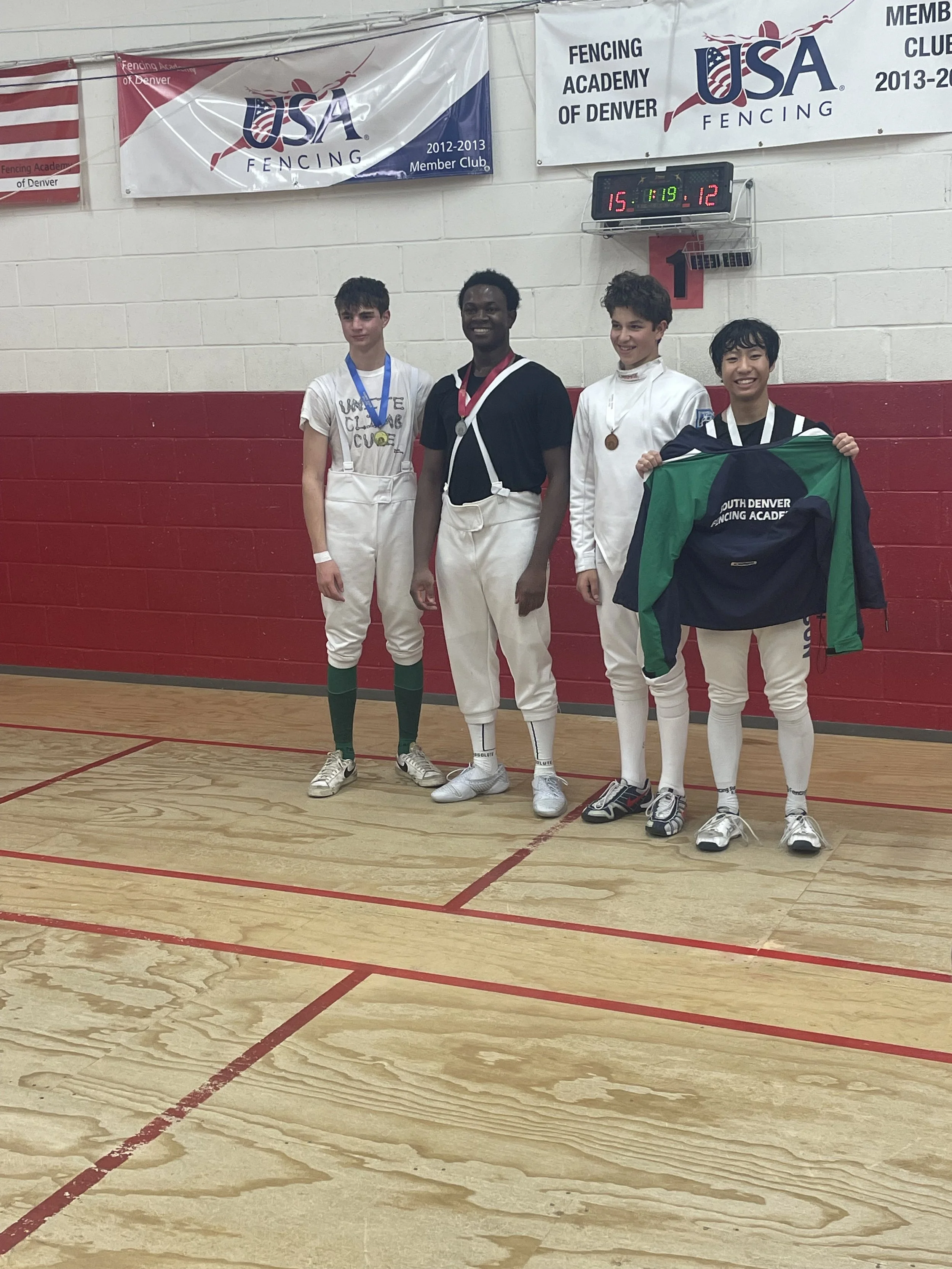 Four young men standing on a wooden sports court, wearing fencing uniforms and medals, with a man holding a flag. Behind them are banners for the USA Fencing Academy of Denver and a scoreboard showing scores and times.