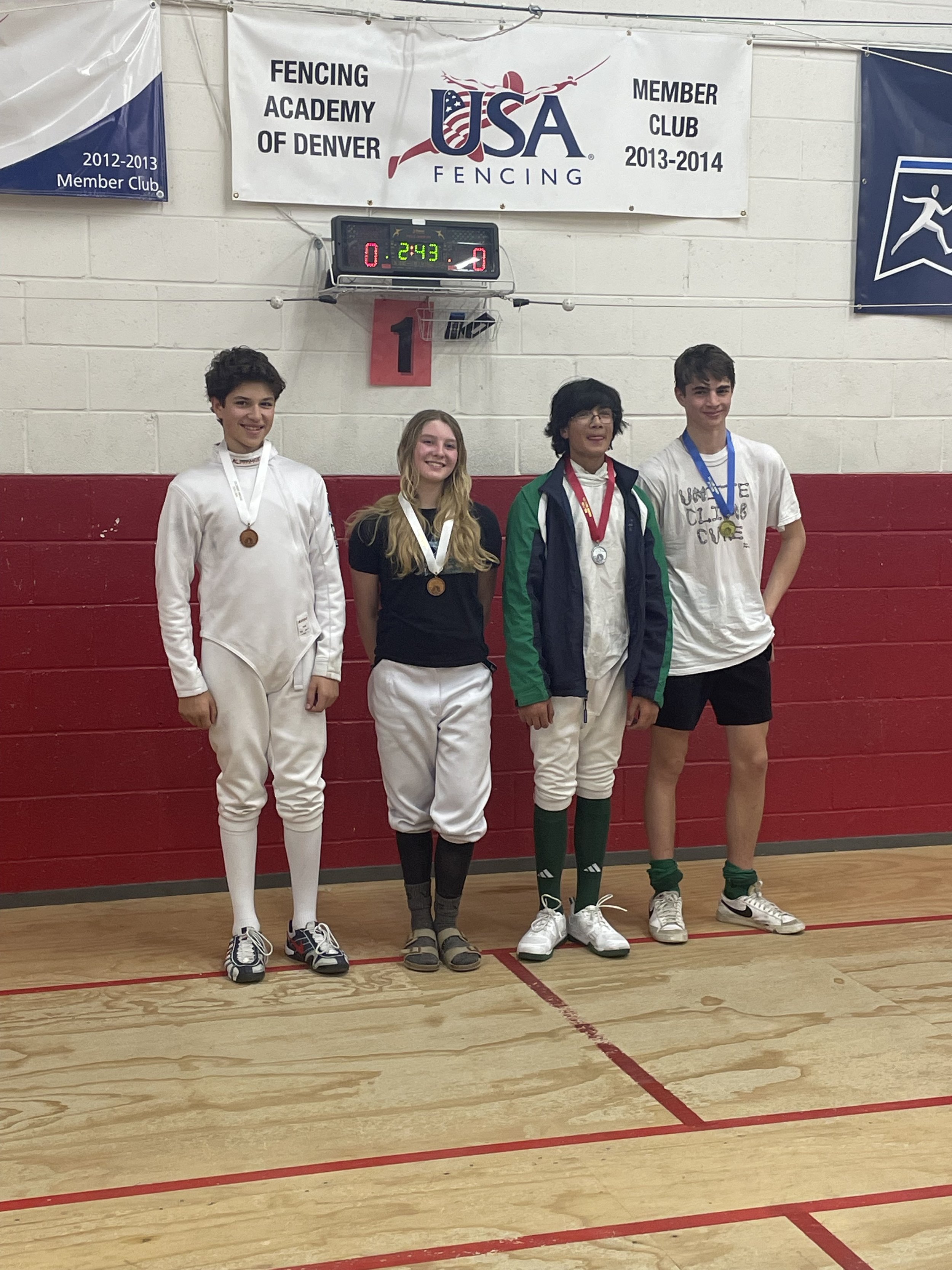 Four young athletes standing on a fencing court, each wearing medal ribbons around their necks; they are smiling after a fencing competition at the USA Fencing Academy of Denver.