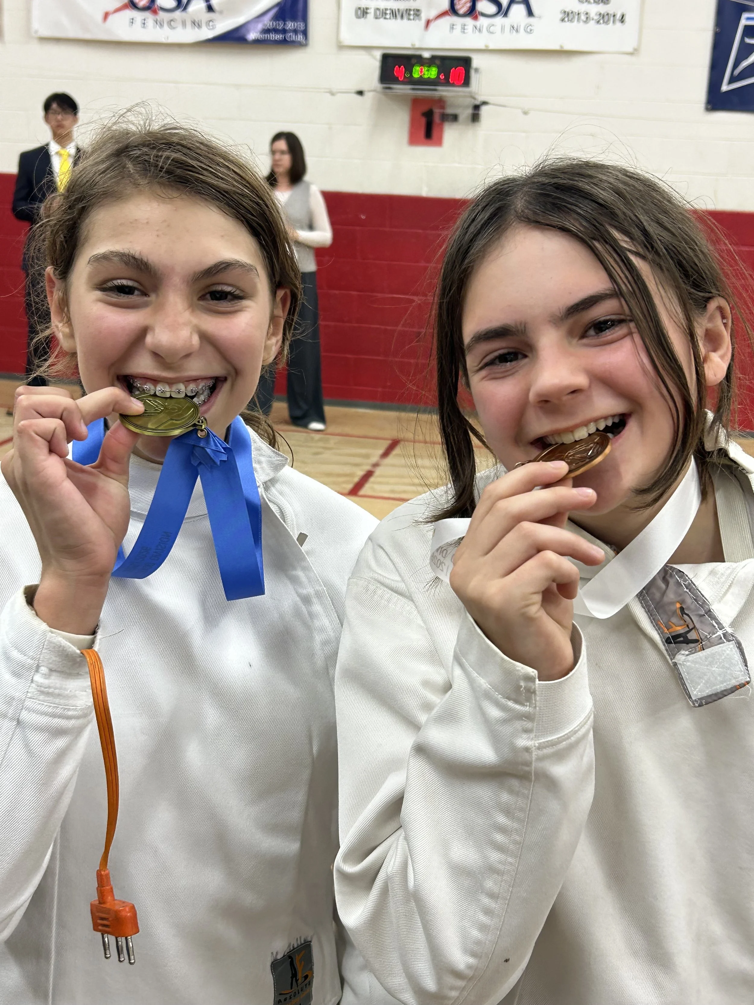 Two young girls in fencing uniforms smiling and holding up medals. They are in a gymnasium with a red and white wall and a scoreboard in the background.