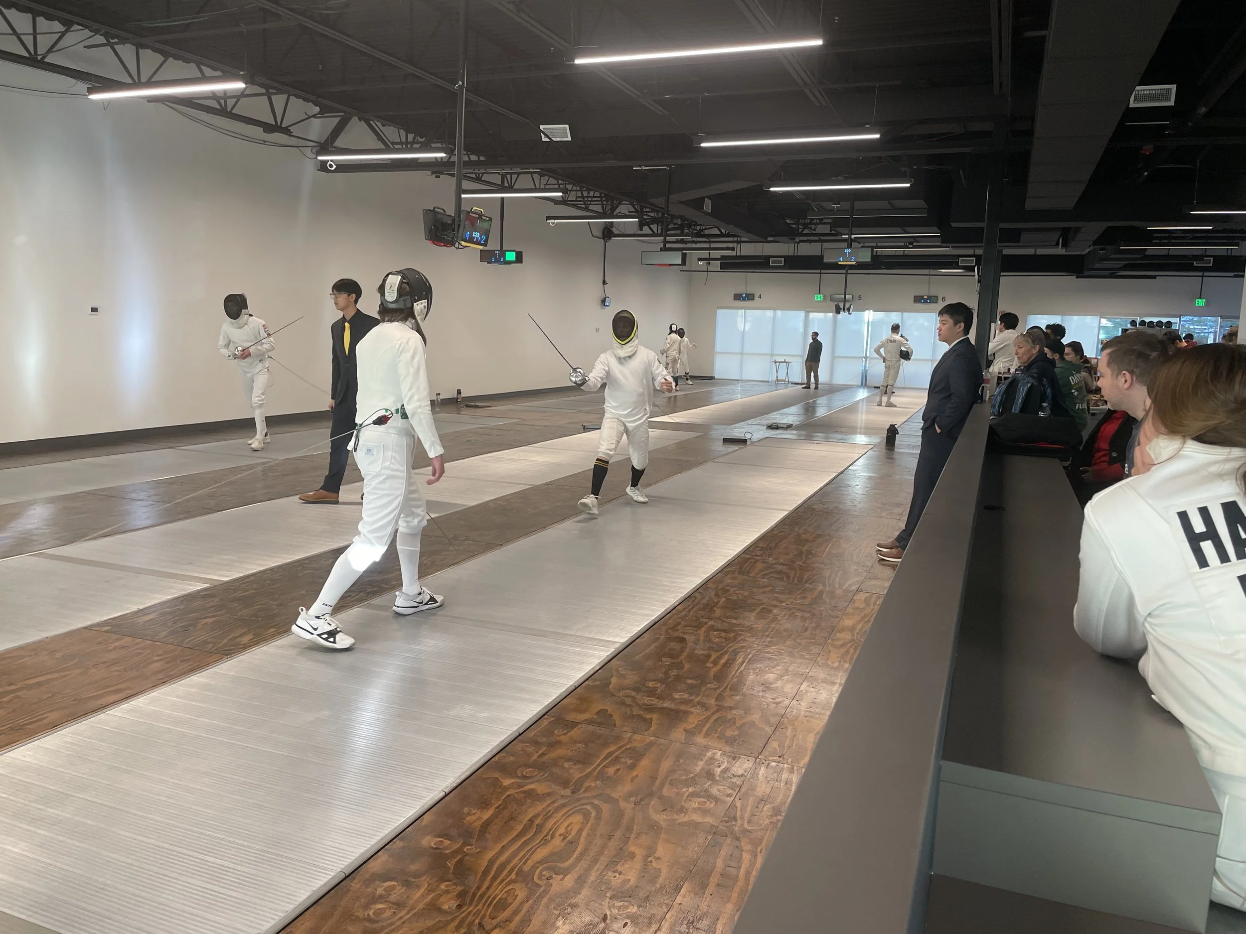 People participating in a fencing competition inside a modern indoor sports facility, with spectators watching from the side.