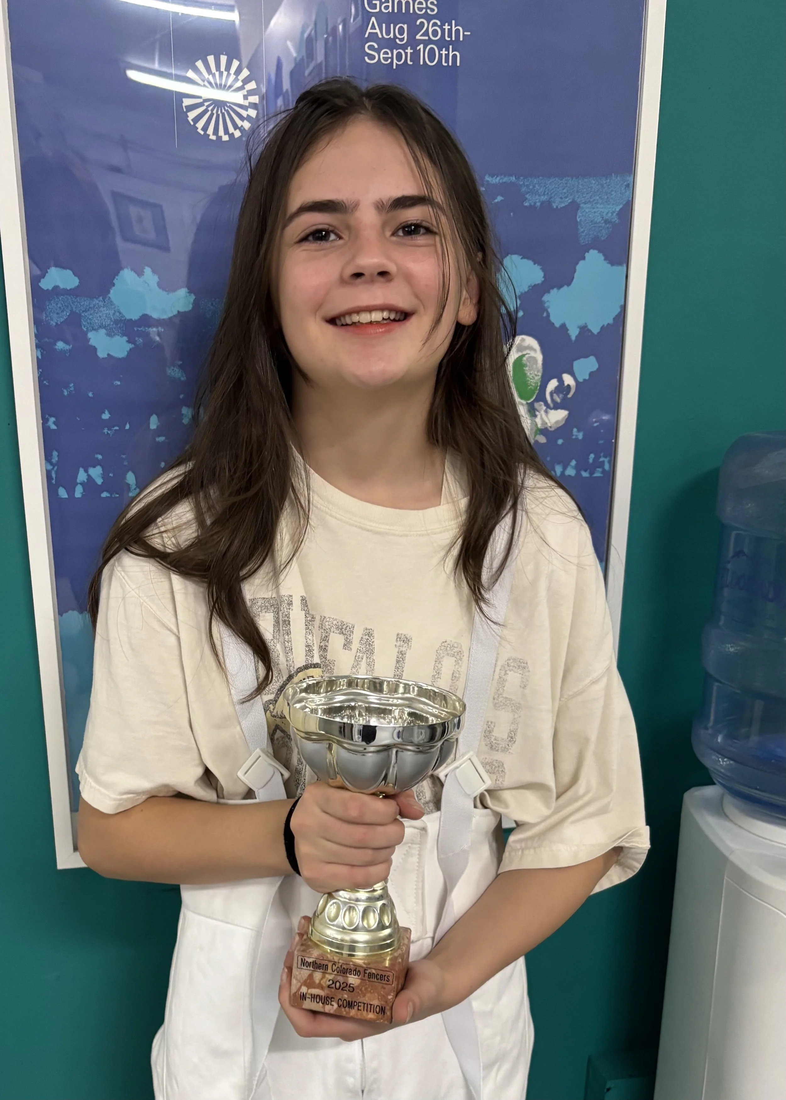 A young girl with long brown hair, smiling, holding a silver trophy with a wooden base, posing in front of a blue poster with event datesYouth fencing in Boulder at Northern Colorado Fencers.