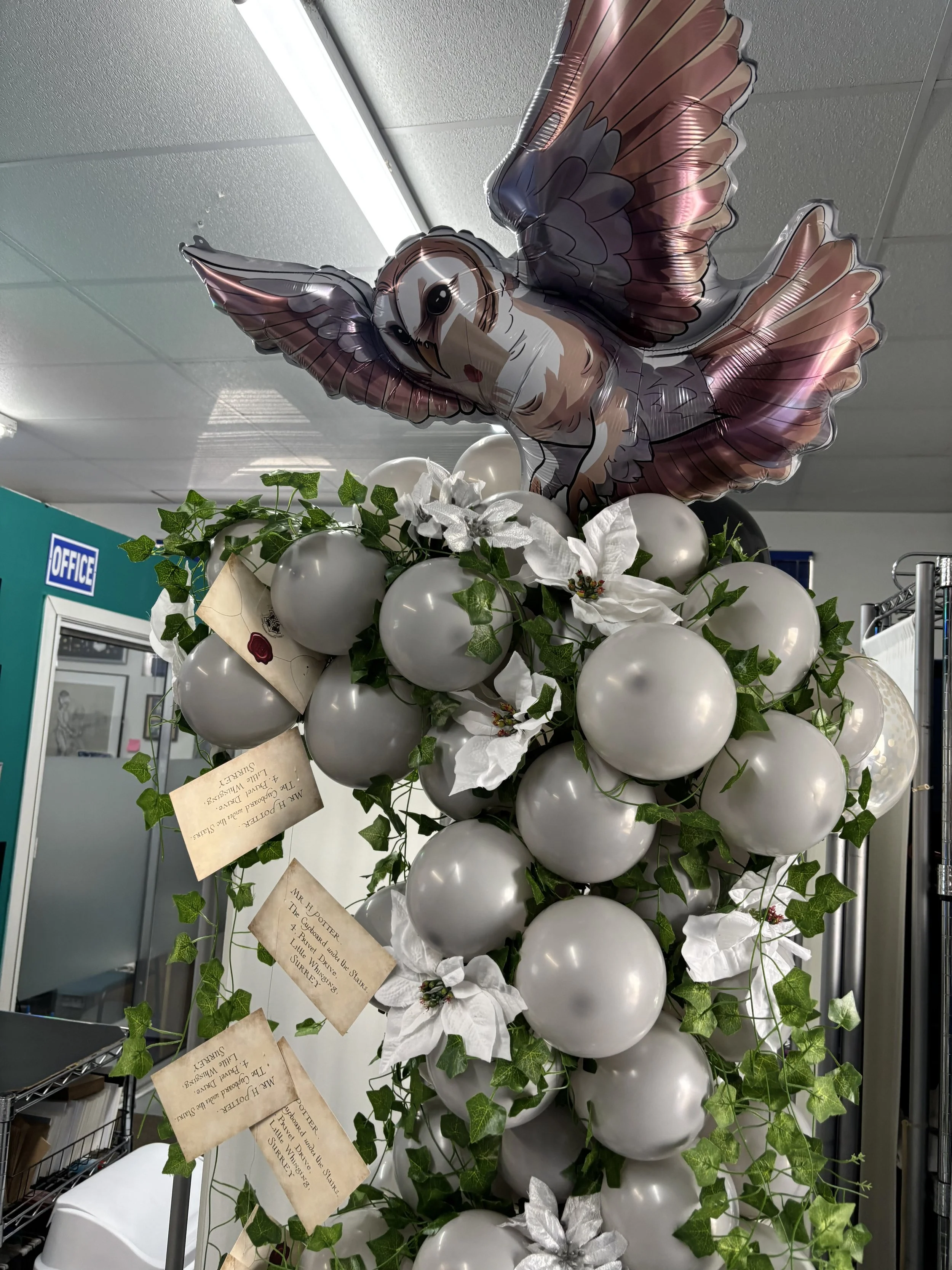 Decorative arrangement with white balloons, artificial white poinsettia flowers, green ivy, and a large owl-shaped foil balloon at the top in a room with white ceiling tiles and an office sign in the background.