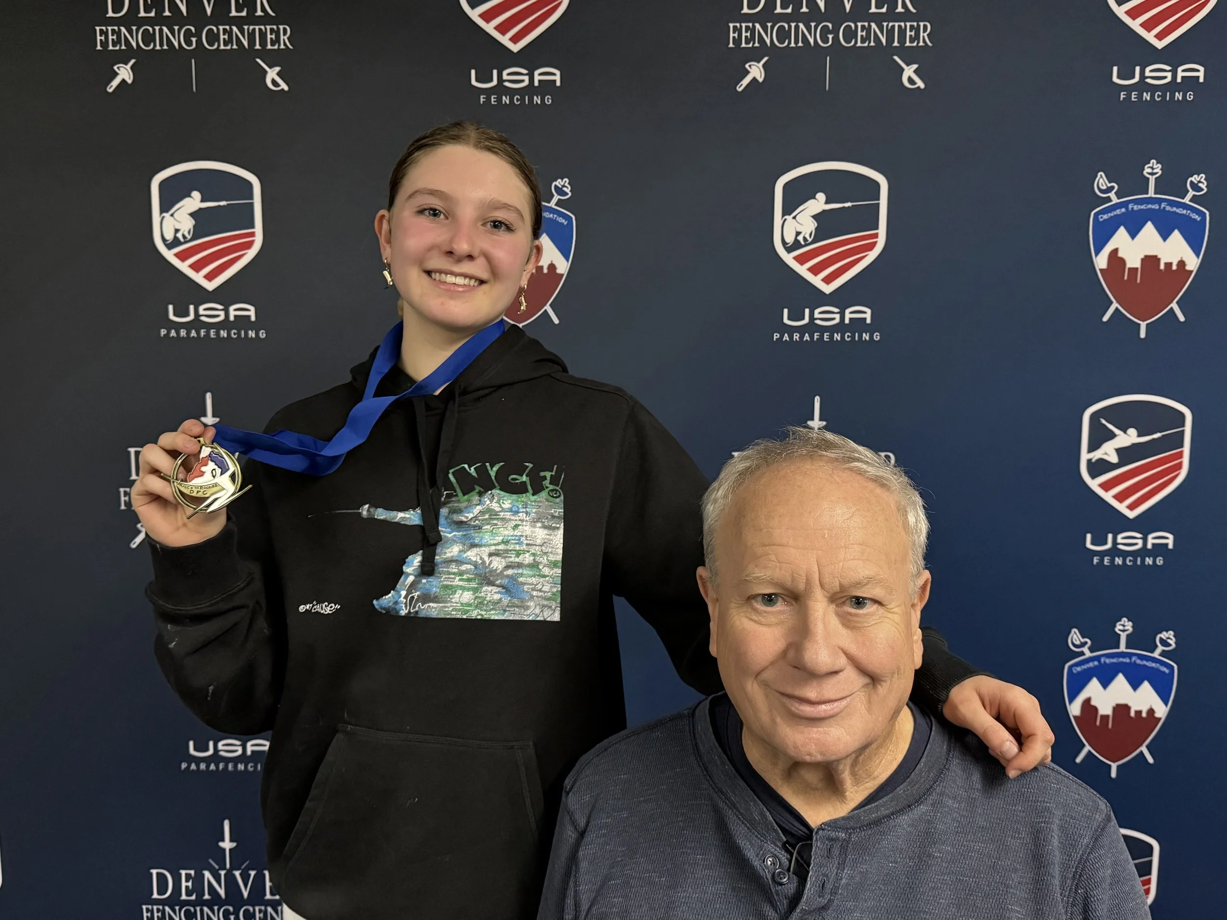 A young woman in a black hoodie holding a medal and smiling, with an older man in front of a backdrop with USA Fencing logos. The young woman has her arm around the man's shoulder at a fencing eventNorthern Colorado Fencers - Fencing, fencing for chi