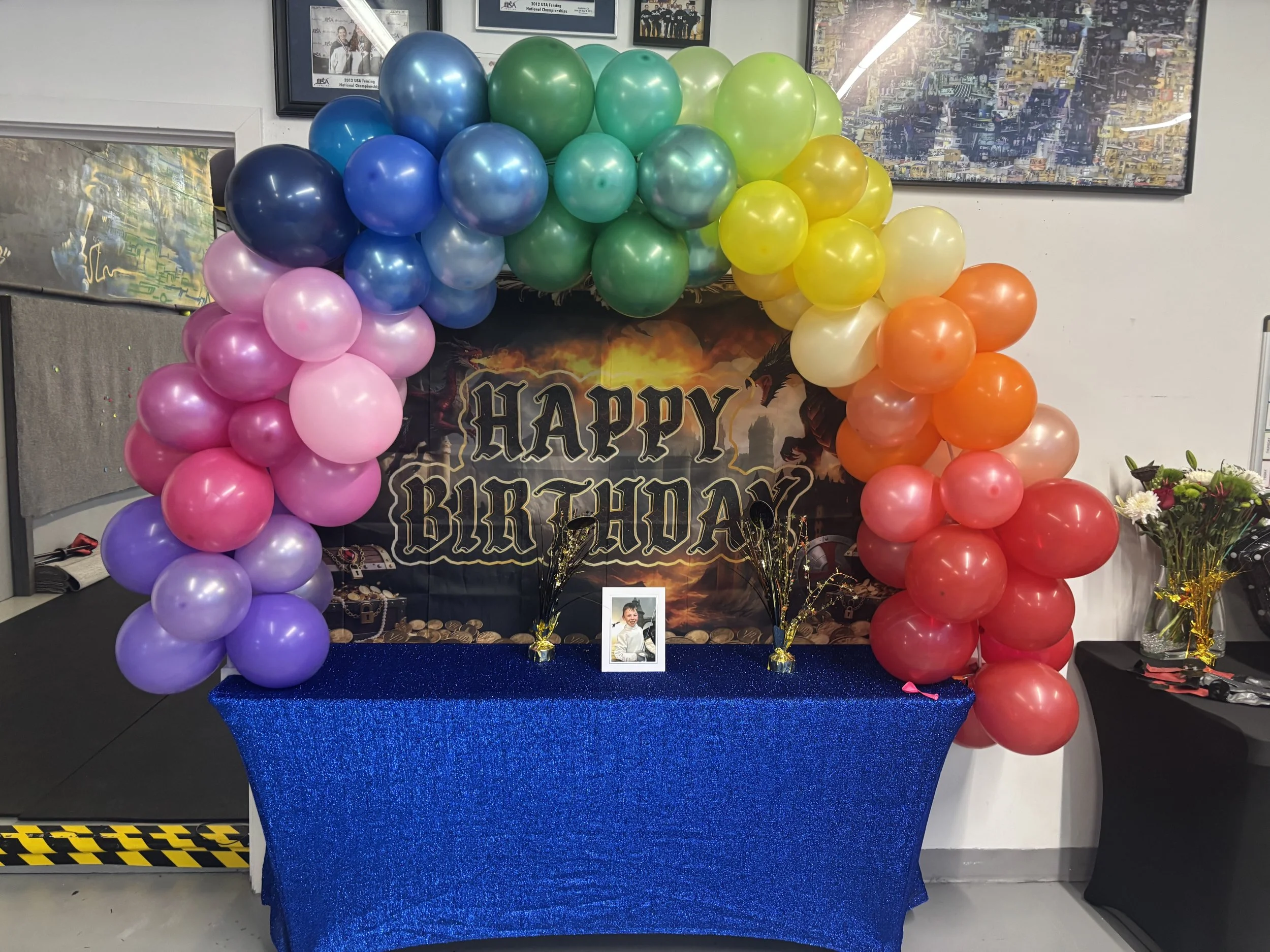 Colorful balloon arch in rainbow colors over a blue tablecloth with a framed photo, birthday message, and party decorations, celebrating a birthday.
