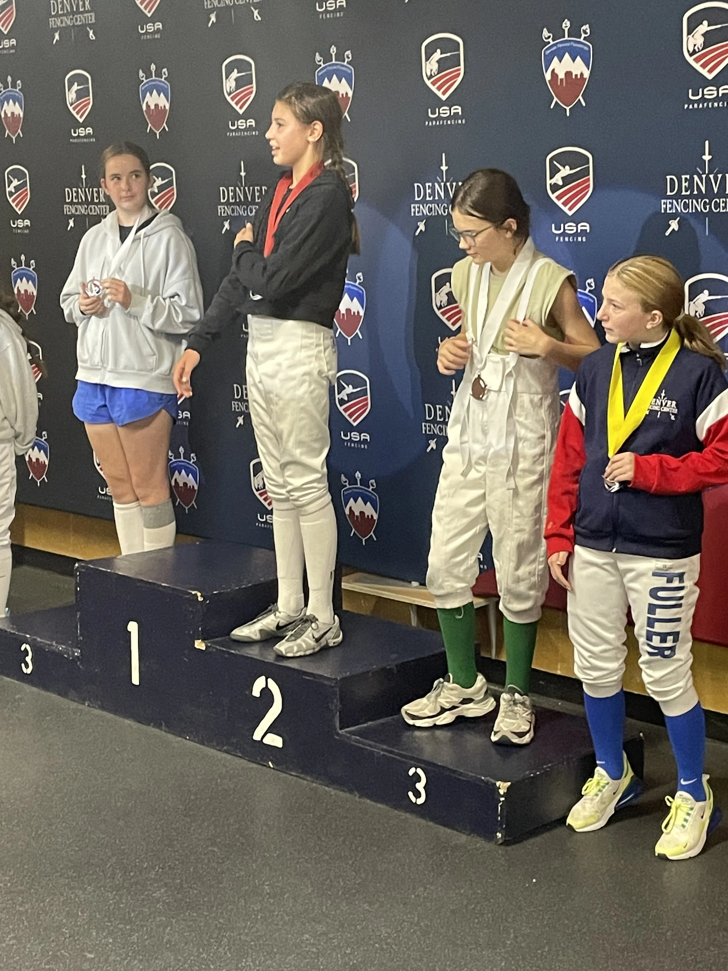 Four young female fencers standing on a winners' podium at a fencing competition award ceremony. The girl in the center is on the top step with a silver medal, the girl on her left is on the second step with a bronze medal, and the girl on the right 