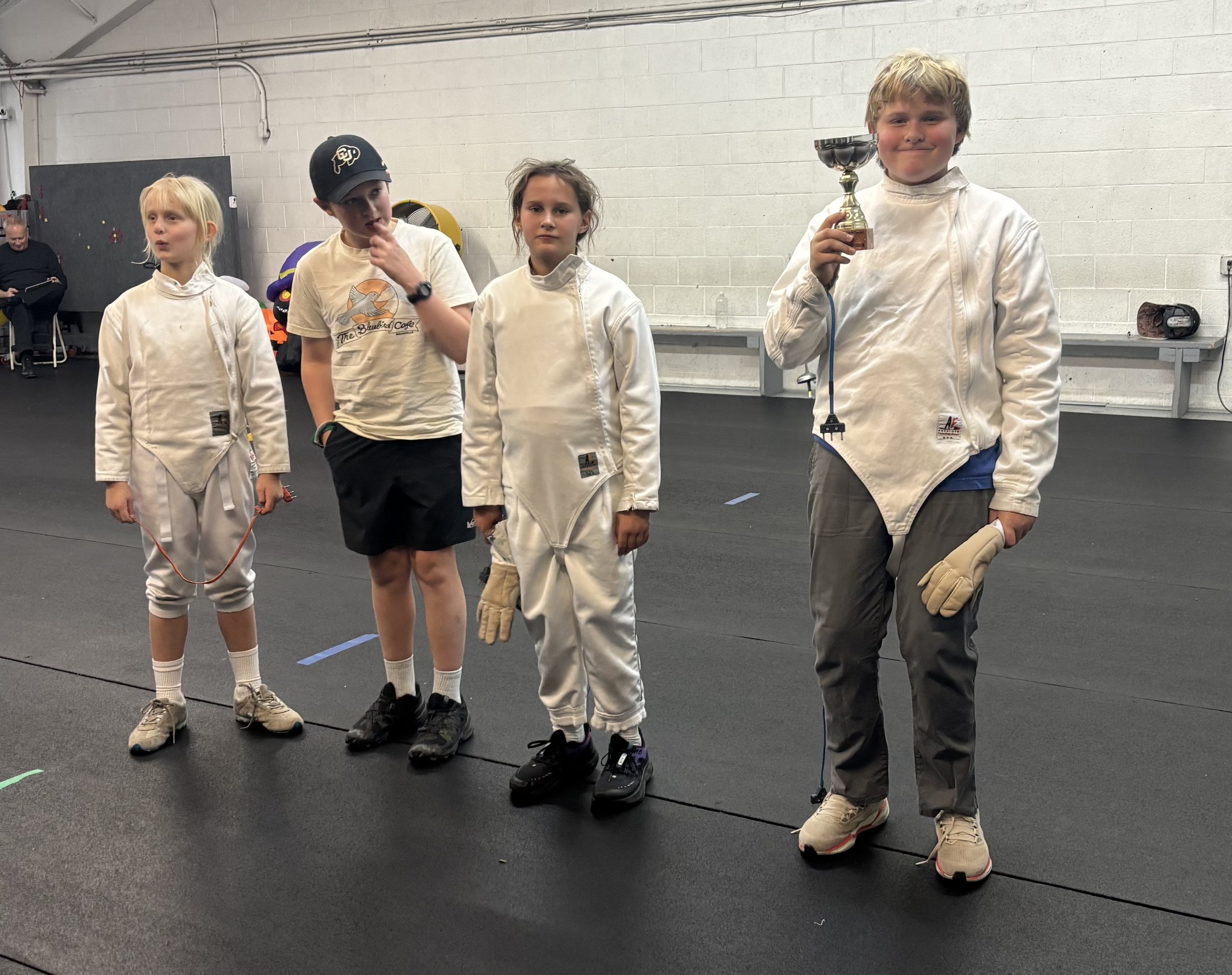 Four young children in fencing attire standing on an indoor fencing strip, with one boy on the far right holding a trophy and smiling at the camera.