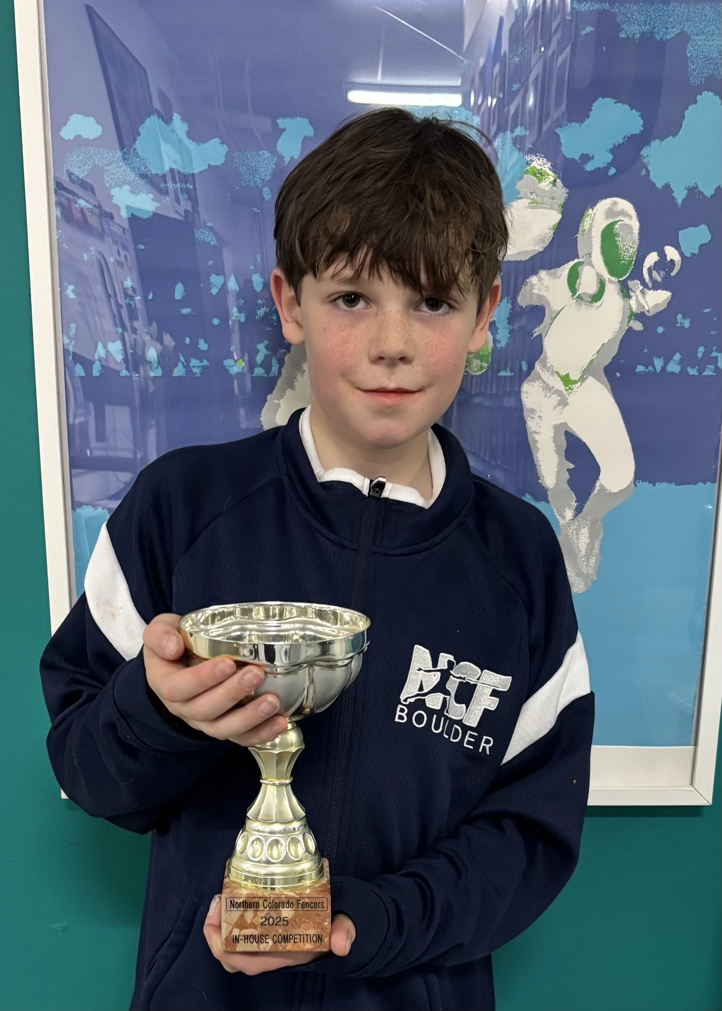 A young boy with brown hair and freckles, wearing a navy blue jacket with white accents, holding a silver trophy with a wooden base that reads 'Northern Colorado Fencers 2025 In-House Competition.' Behind him is a blue background featuring a stylized