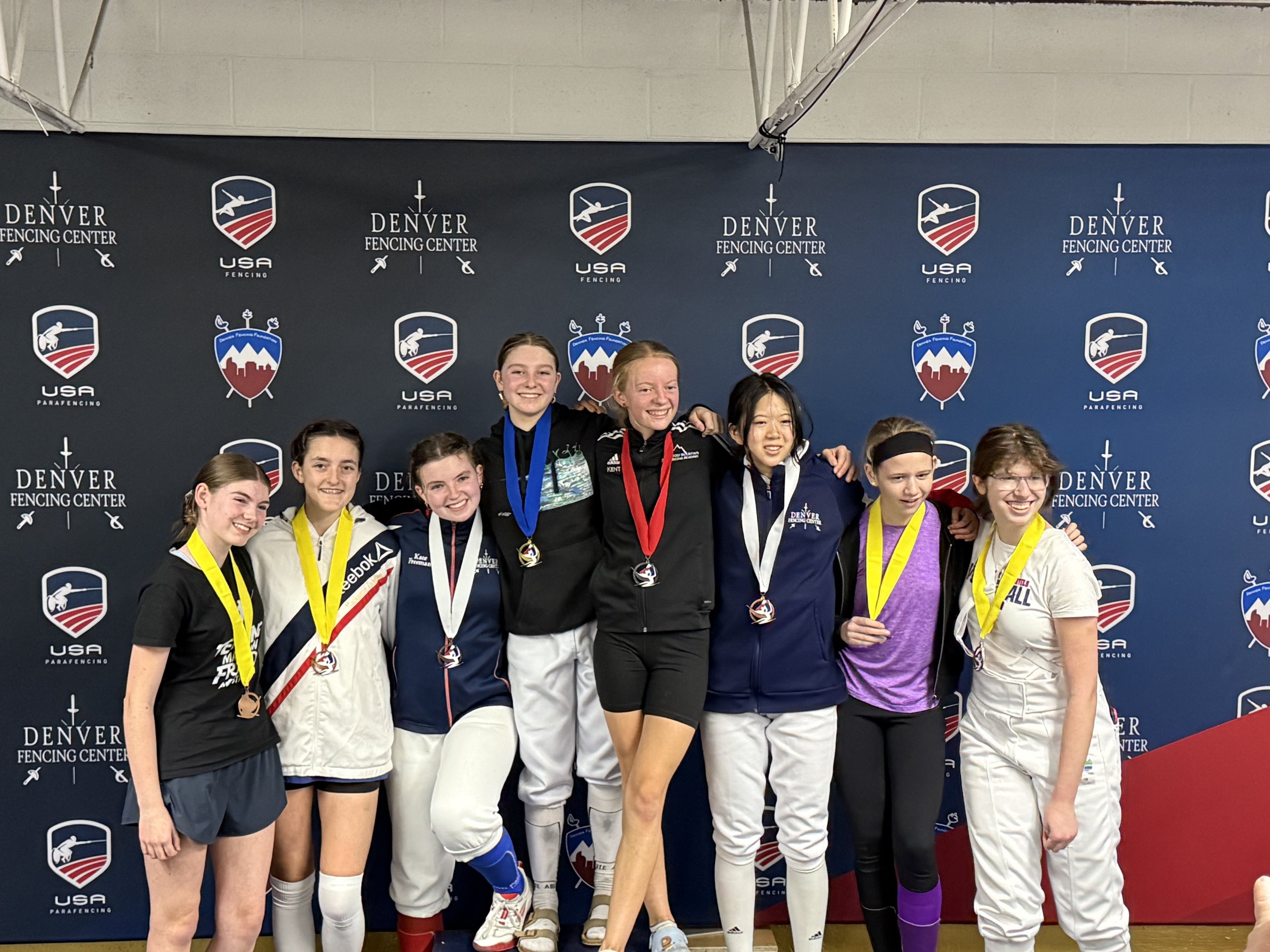 Group of eight young female athletes standing in front of a backdrop with Denver Fencing Center and USA Fencing logos, some wearing medals around their necks, celebrating after a competition.
