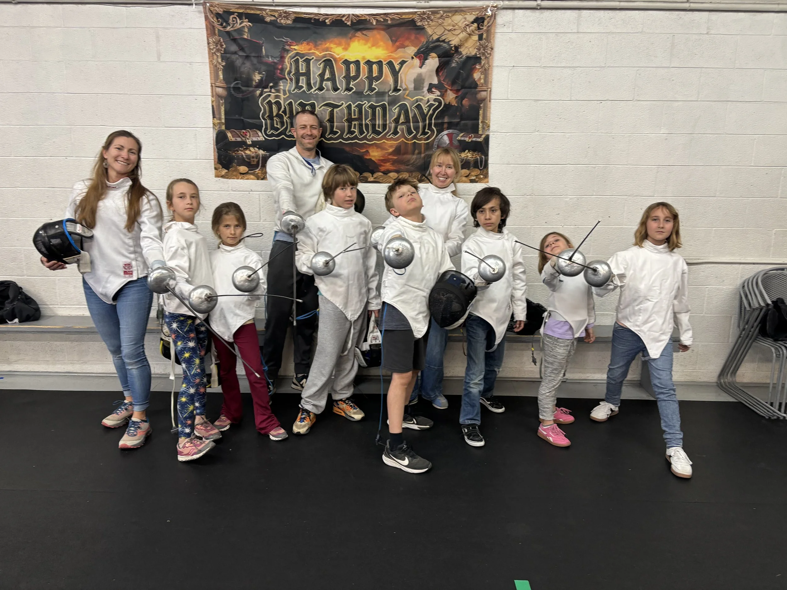 Group of children and two adults dressed in fencing gear, standing in front of a birthday banner that says 'Happy Birthday' with a dragon and treasure chest decorations. Boulder birthday party.