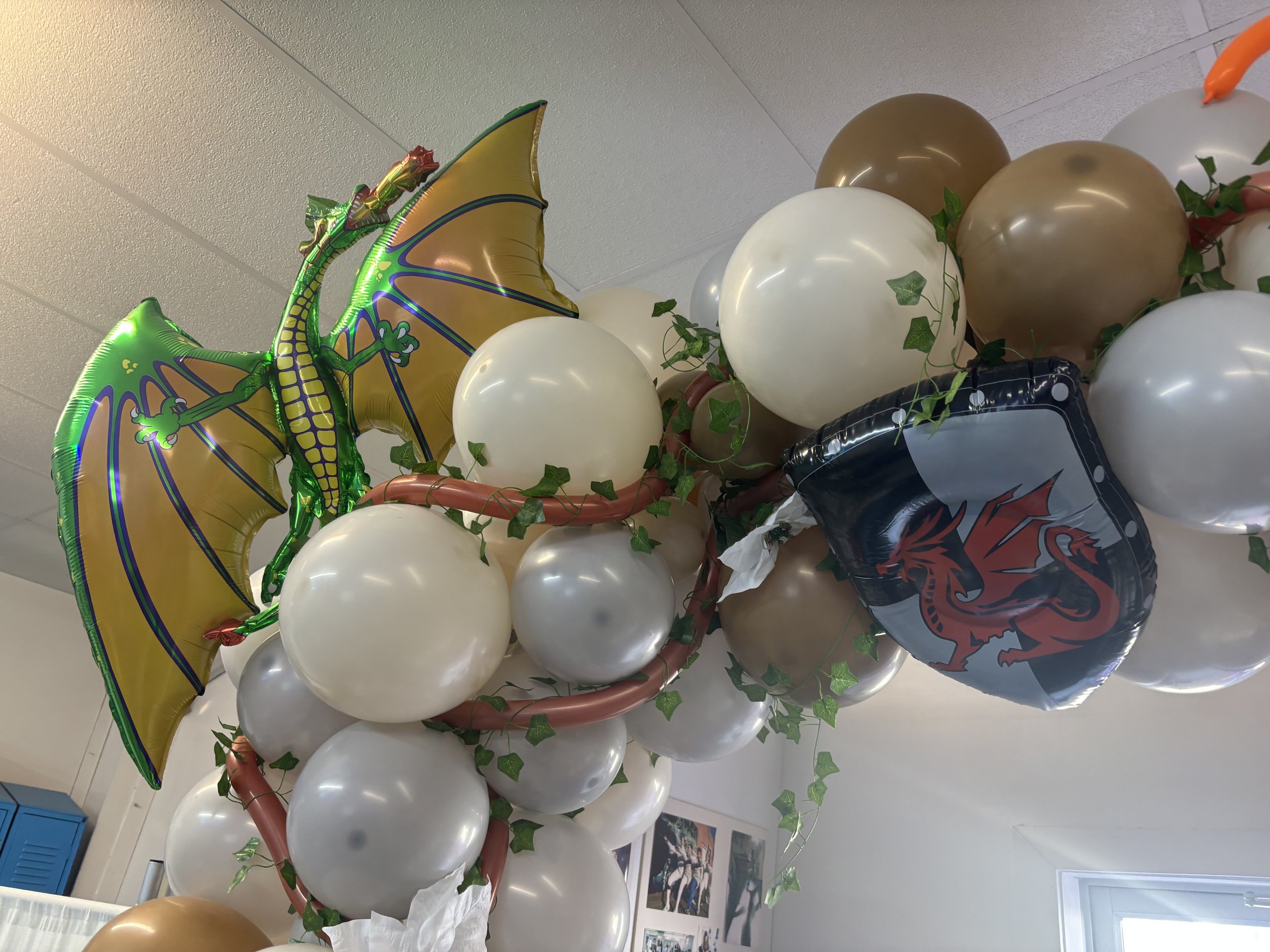Decorative arrangement of white, brown, and silver balloons with green Ivy leaves, featuring a large yellow and green dragon balloon with wings and a black and red dragon balloon.