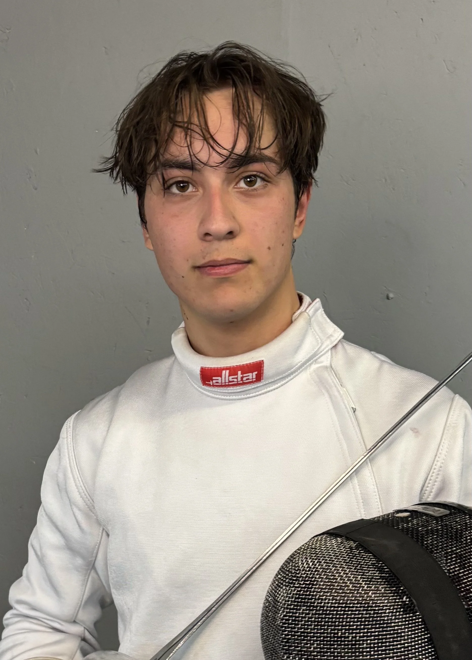 Young man wearing a white fencing jacket with the brand 'allstar' on the collar, holding a fencing mask and foil, standing against a gray wall.Youth fencing in Boulder at Northern Colorado Fencers.