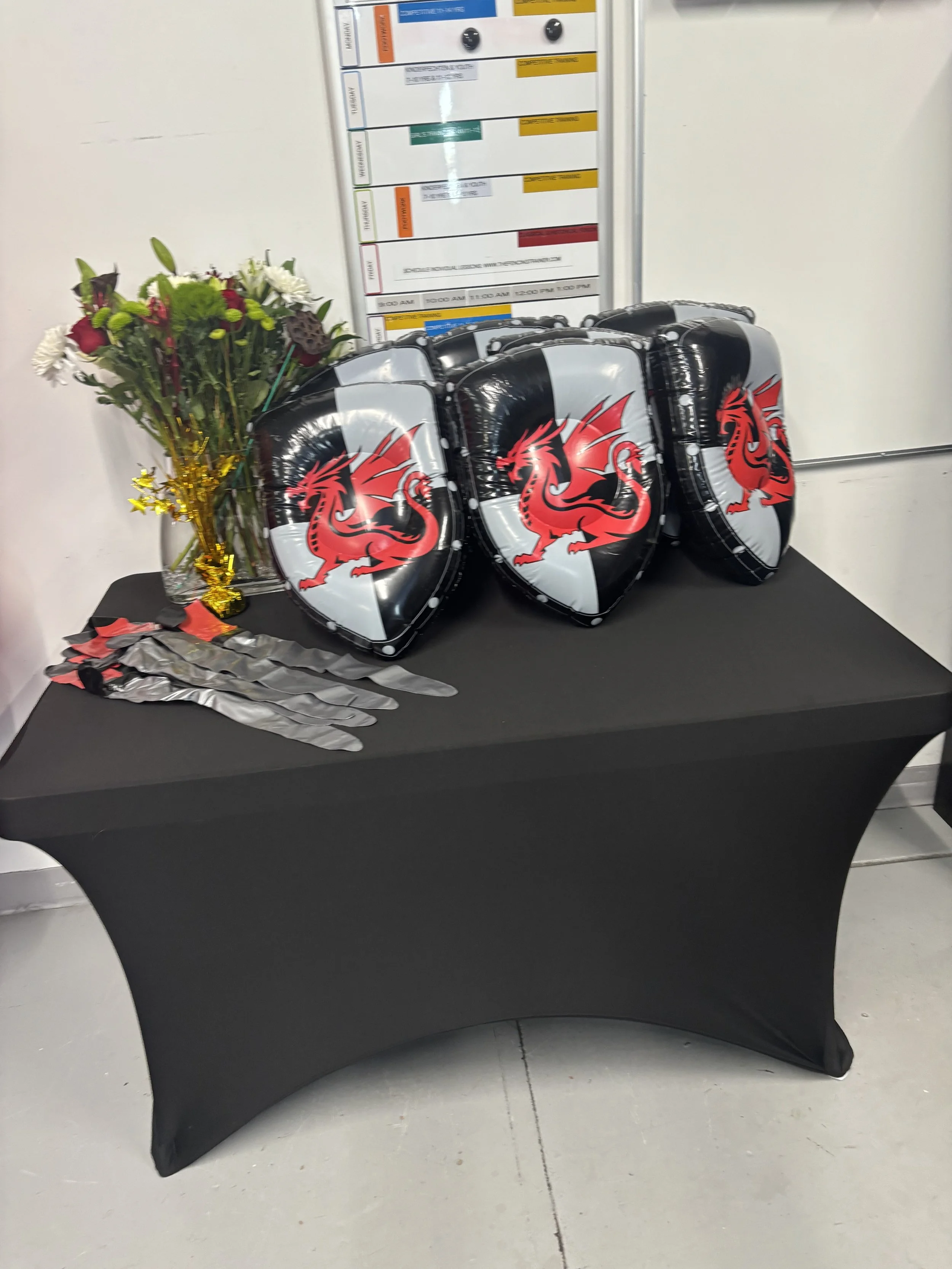 Table with three inflatable shields featuring a red and black dragon emblem, a bouquet of flowers, and some model skeleton hands, with a whiteboard and schedule in the background.