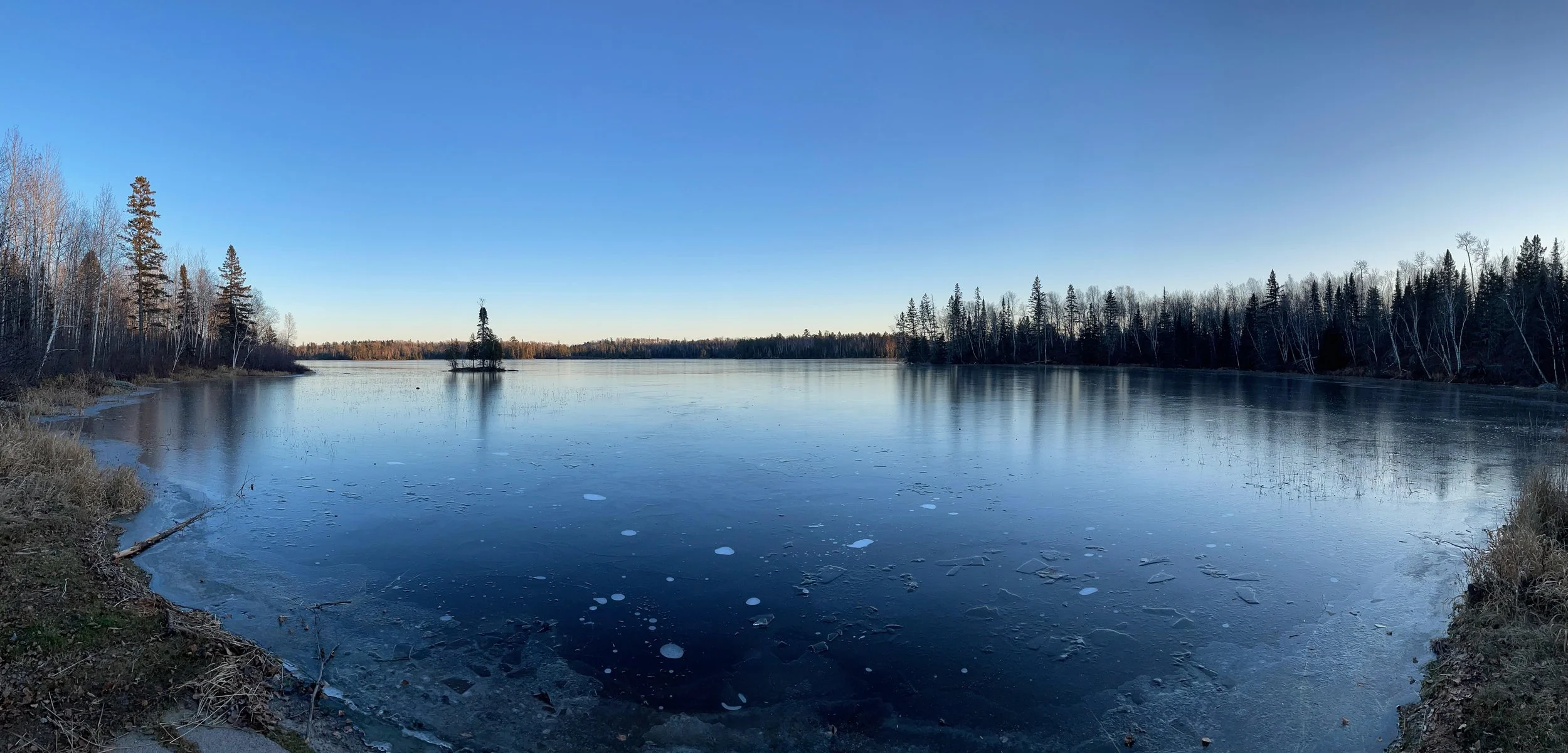 Panoramic early frozen lake with island under blue sky in Ely MN, BWCA area, December 5 2023. Arrowhead Outdoors Ely MN winter fishing report featuring ice house rentals, premium tackle, hunting gear & camping supplies.