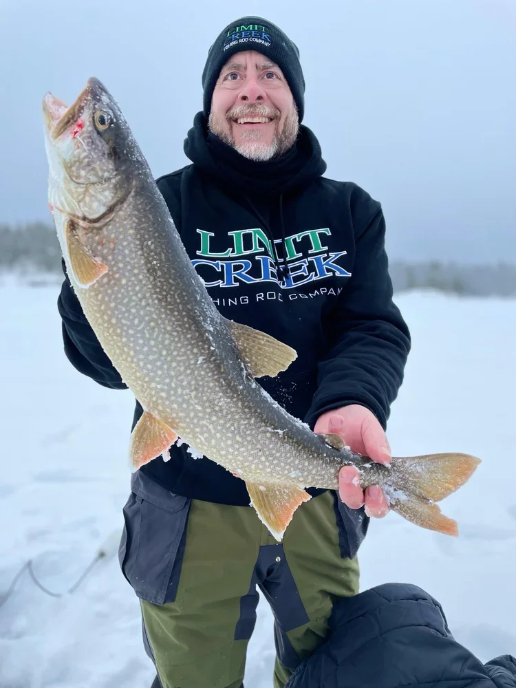 Ely MN BWCA lake trout catch in Arrowhead Outdoors fishing report January 4 2023 – angler holding spotted laker on snowy ice with winter BWCA backdrop