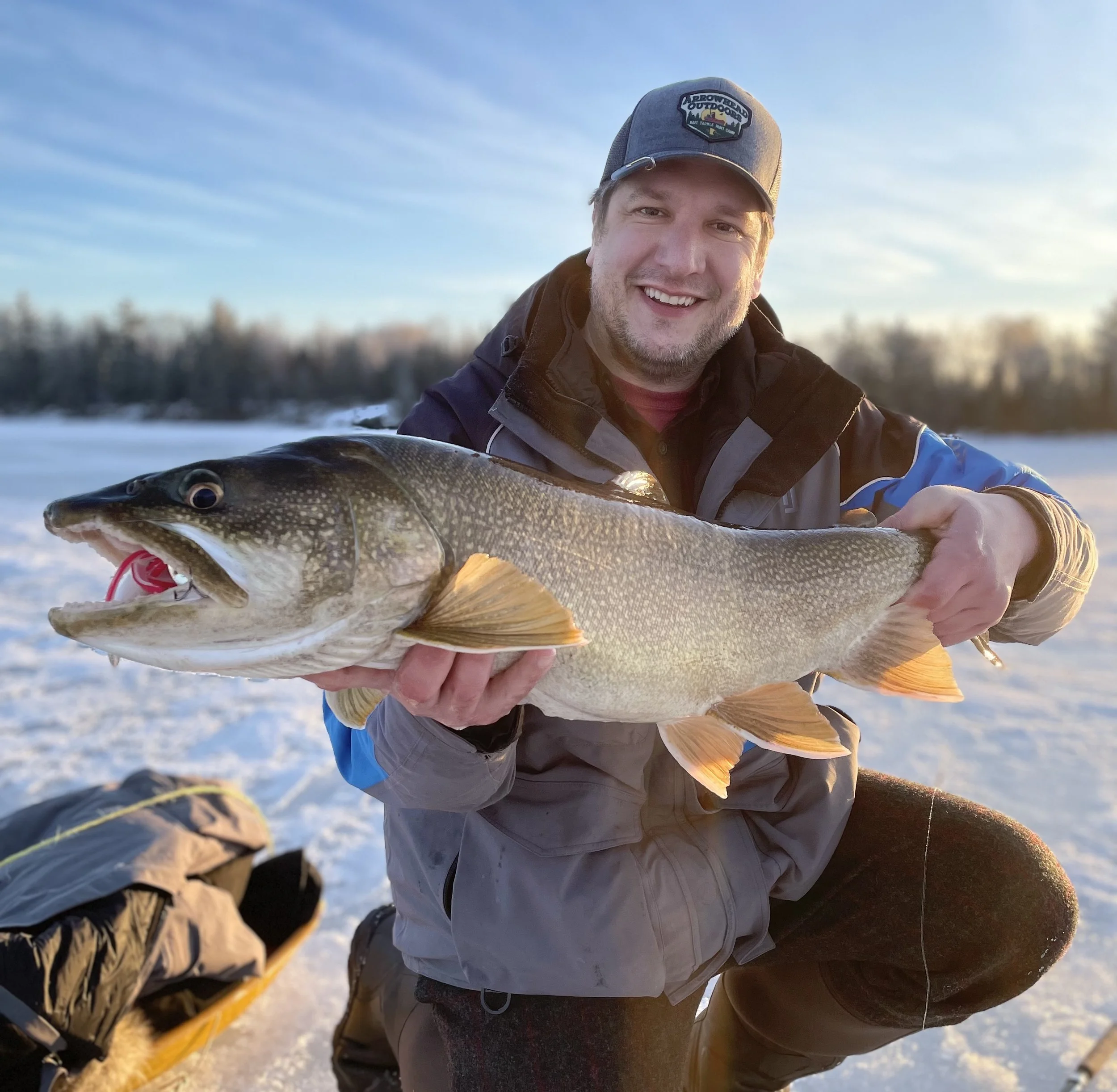 Angler holds lake trout caught on Laker Tackle tube on snowy ice in Ely MN, BWCA area, February 6 2024. Arrowhead Outdoors Ely MN winter fishing report with ice house rentals, premium tackle, hunting gear & camping supplies.
