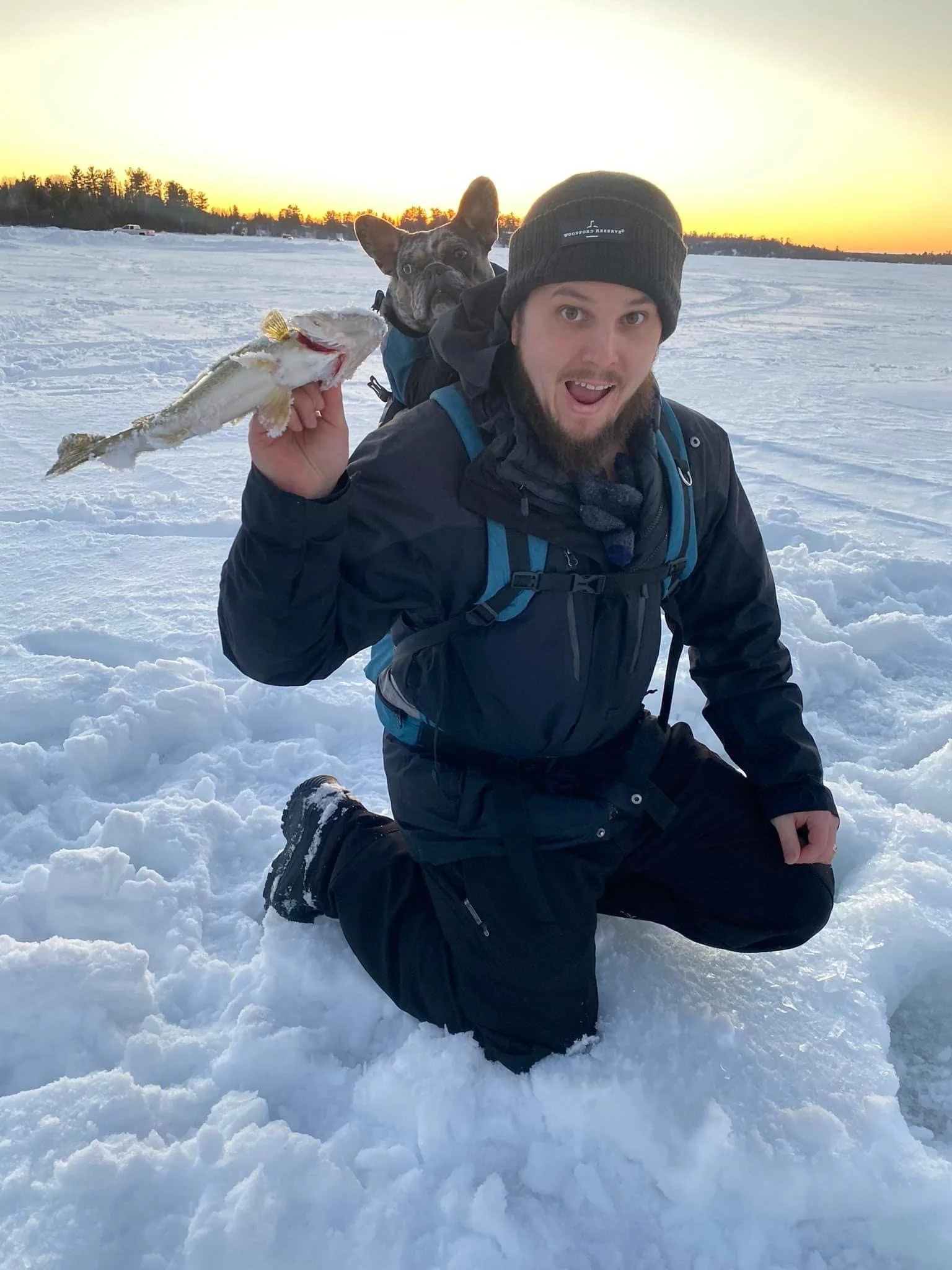 Ely MN BWCA walleye catch in Arrowhead Outdoors fishing report February 21 2023 – angler kneeling on ice holding walleye with dog in backpack at sunset