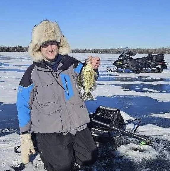Angler holds crappie on snowy ice with snowmobile in Ely MN, BWCA area, January 2 2024. Arrowhead Outdoors Ely MN winter fishing report with ice house rentals, premium tackle, hunting gear & camping supplies.
