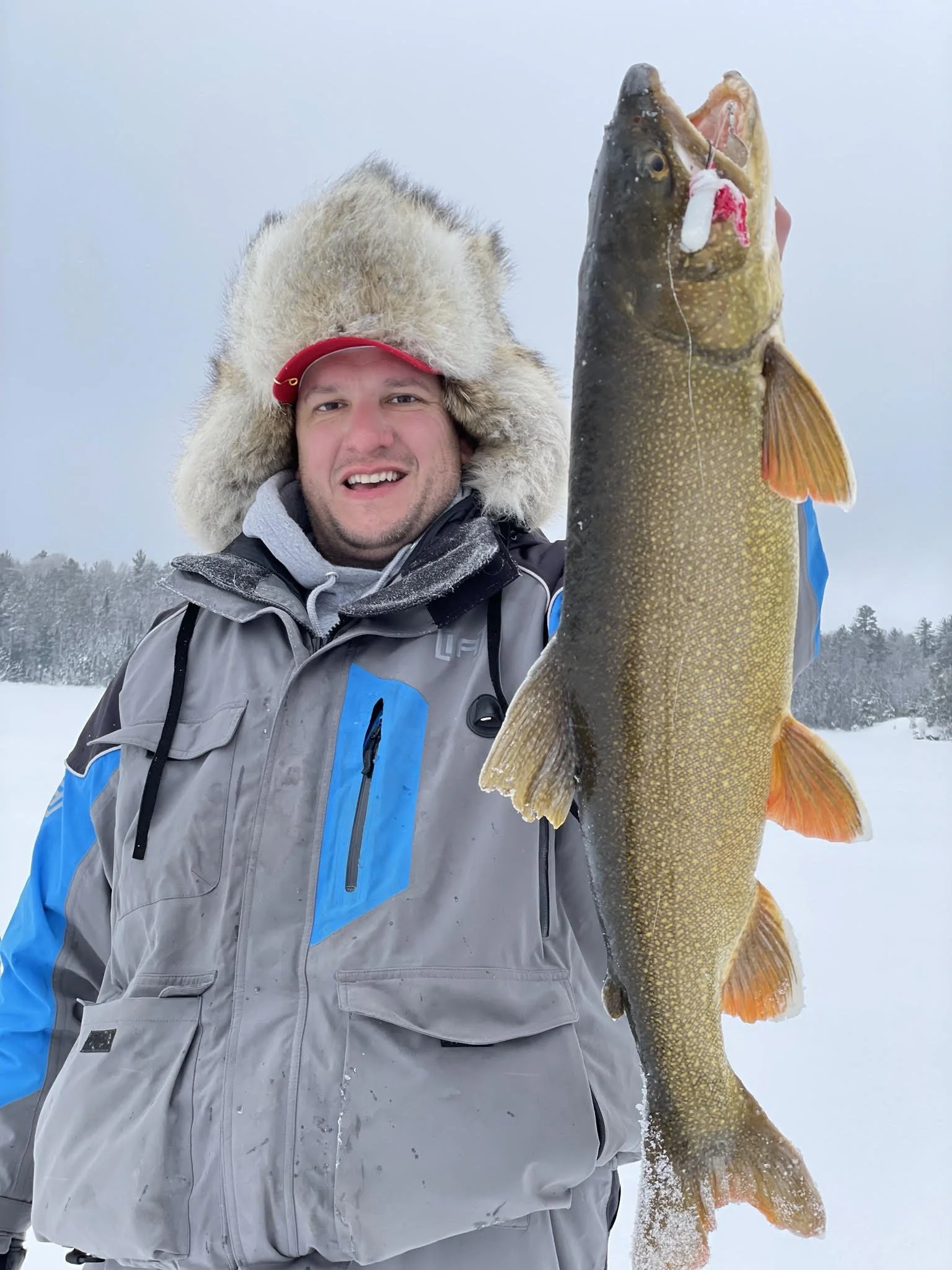Angler holds lake trout caught on Laker Tackle tube on snowy ice in Ely MN, BWCA area, January 9 2024. Arrowhead Outdoors Ely MN winter fishing report with ice house rentals, premium tackle, hunting gear & camping supplies.