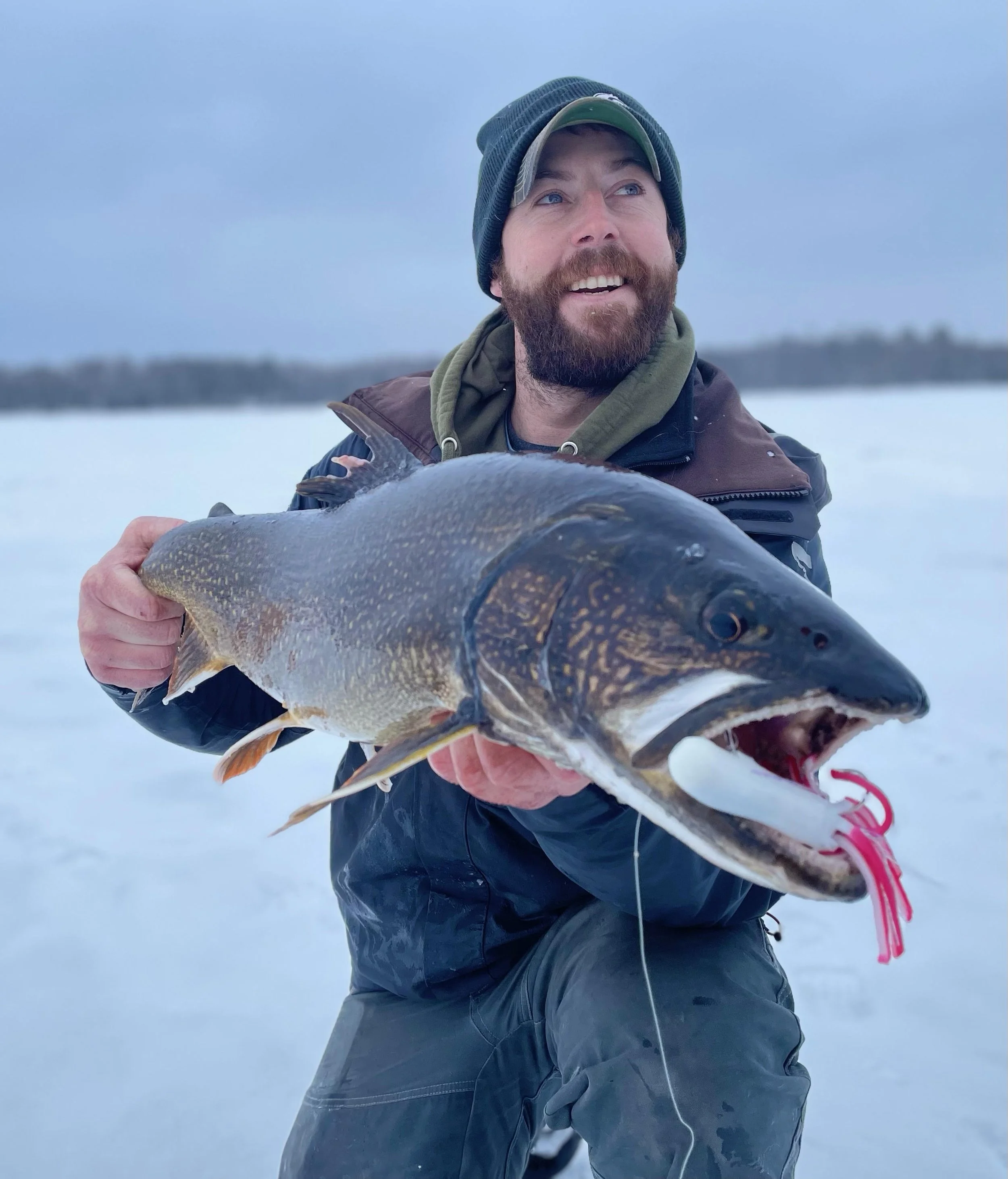 Angler holds lake trout caught on Laker Tackle tube on snowy ice in Ely MN, BWCA area, January 23 2024. Arrowhead Outdoors Ely MN winter fishing report with ice house rentals, premium tackle, hunting gear & camping supplies.