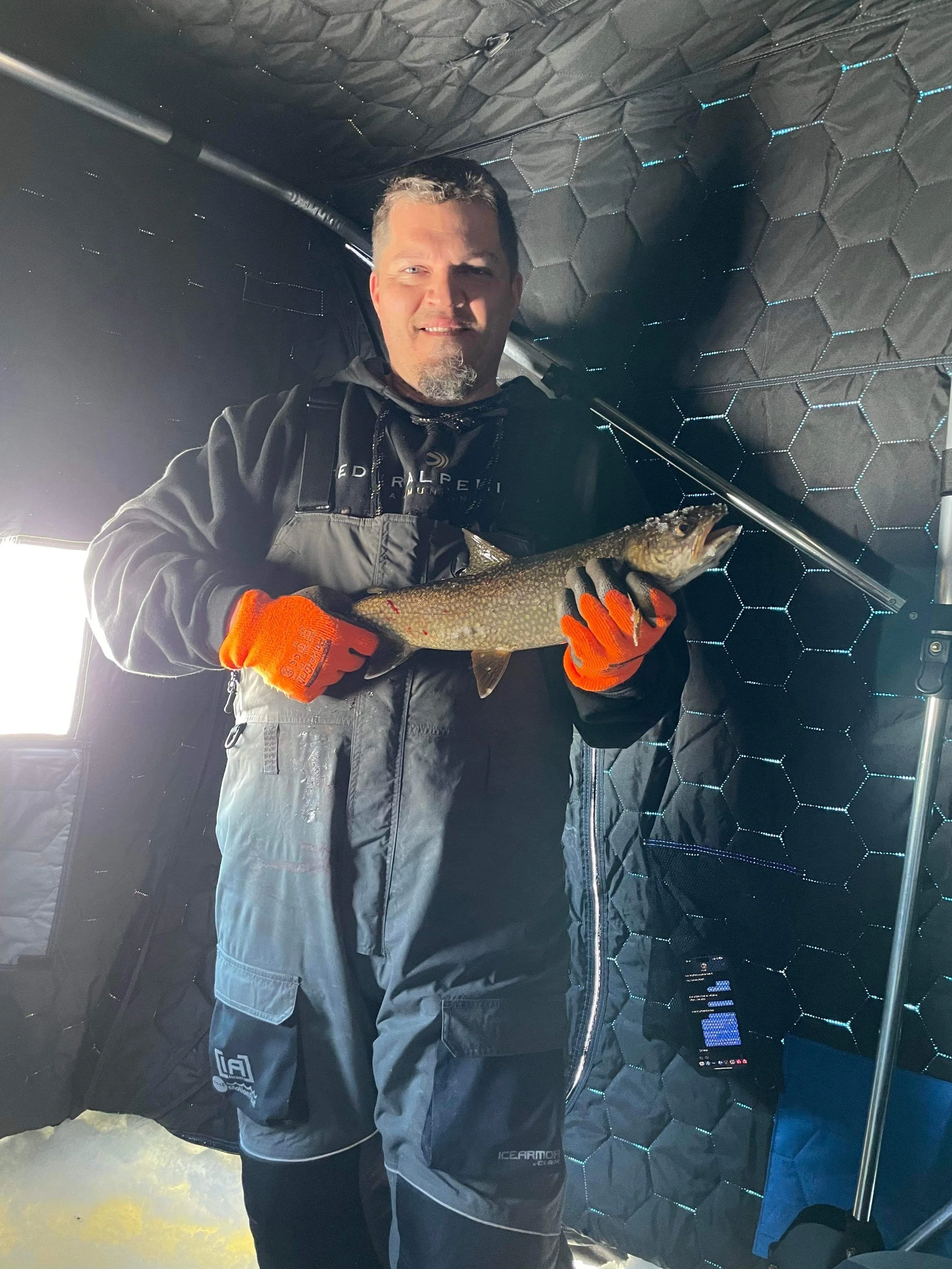 Ely MN BWCA lake trout catch in Arrowhead Outdoors fishing report March 7 2023 – angler inside heated ice house holding spotted laker