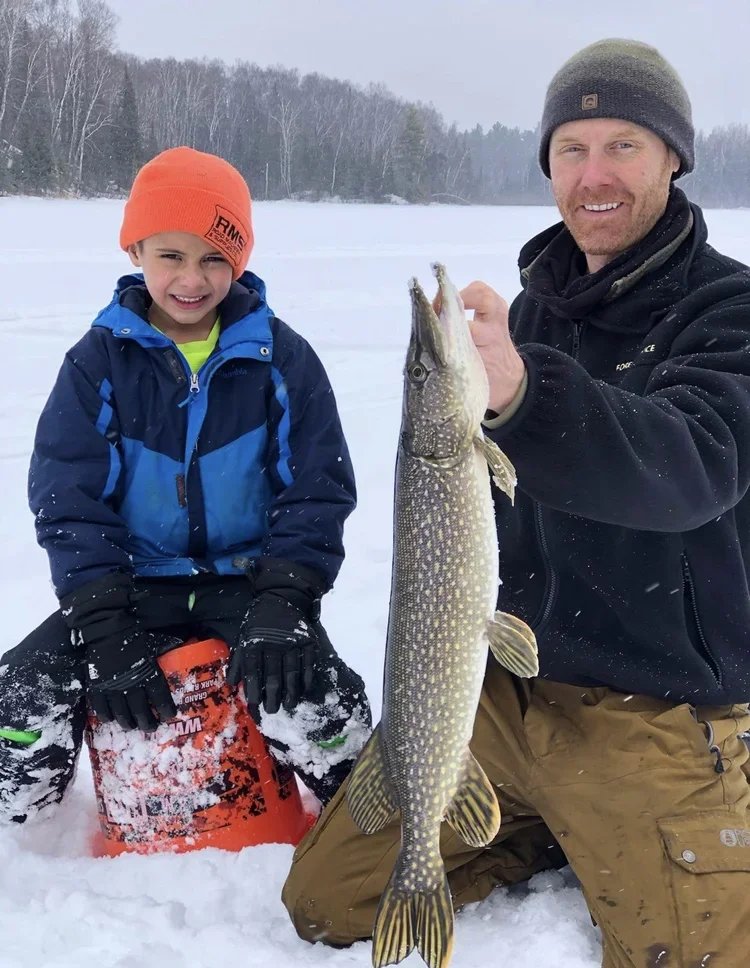 Ely MN BWCA northern pike catch in Arrowhead Outdoors fishing report December 7 2022 – dad and son smiling on ice holding big toothy pike with snowy BWCA backdrop