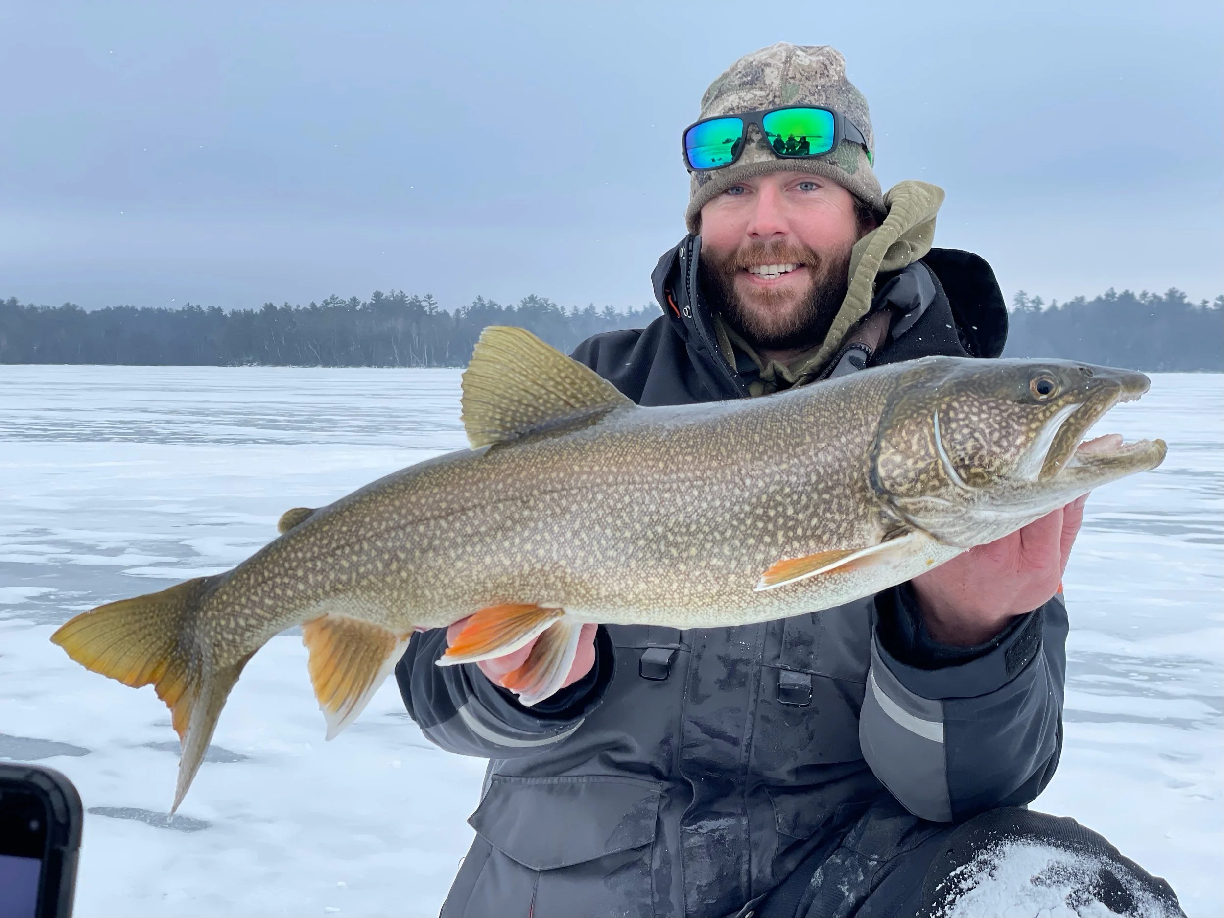 Angler holds lake trout caught on Laker Tackle tube on snowy ice in Ely MN, BWCA area, February 13 2024. Arrowhead Outdoors Ely MN winter fishing report with ice house rentals, premium tackle, hunting gear & camping supplies.
