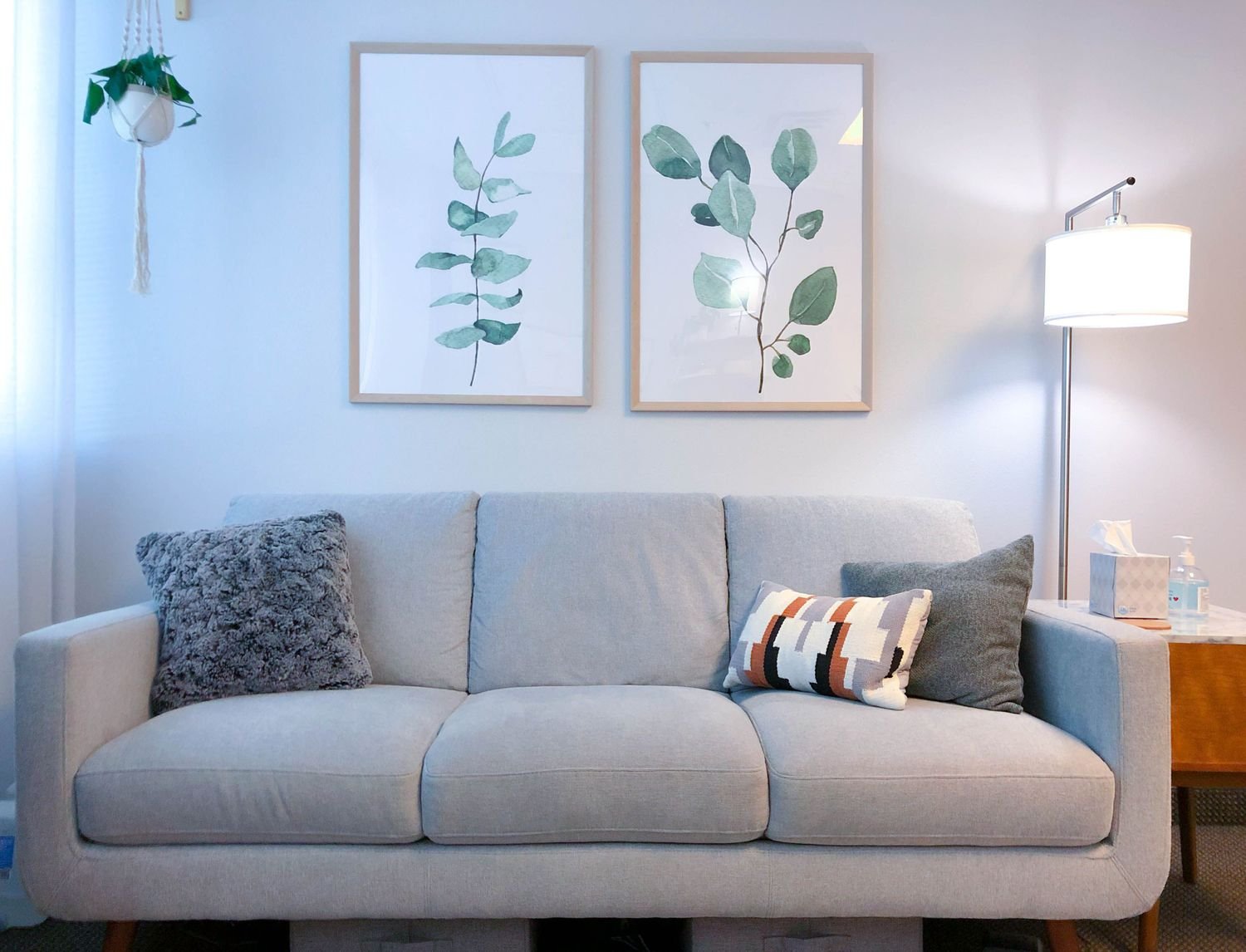 A modern living room with a light gray sofa, decorated with textured and patterned pillows. Two framed botanical prints hang above the sofa. A floor lamp with a white shade is on the right, beside a wooden side table with a tissue box and hand sanitizer. A hanging plant is on the left.