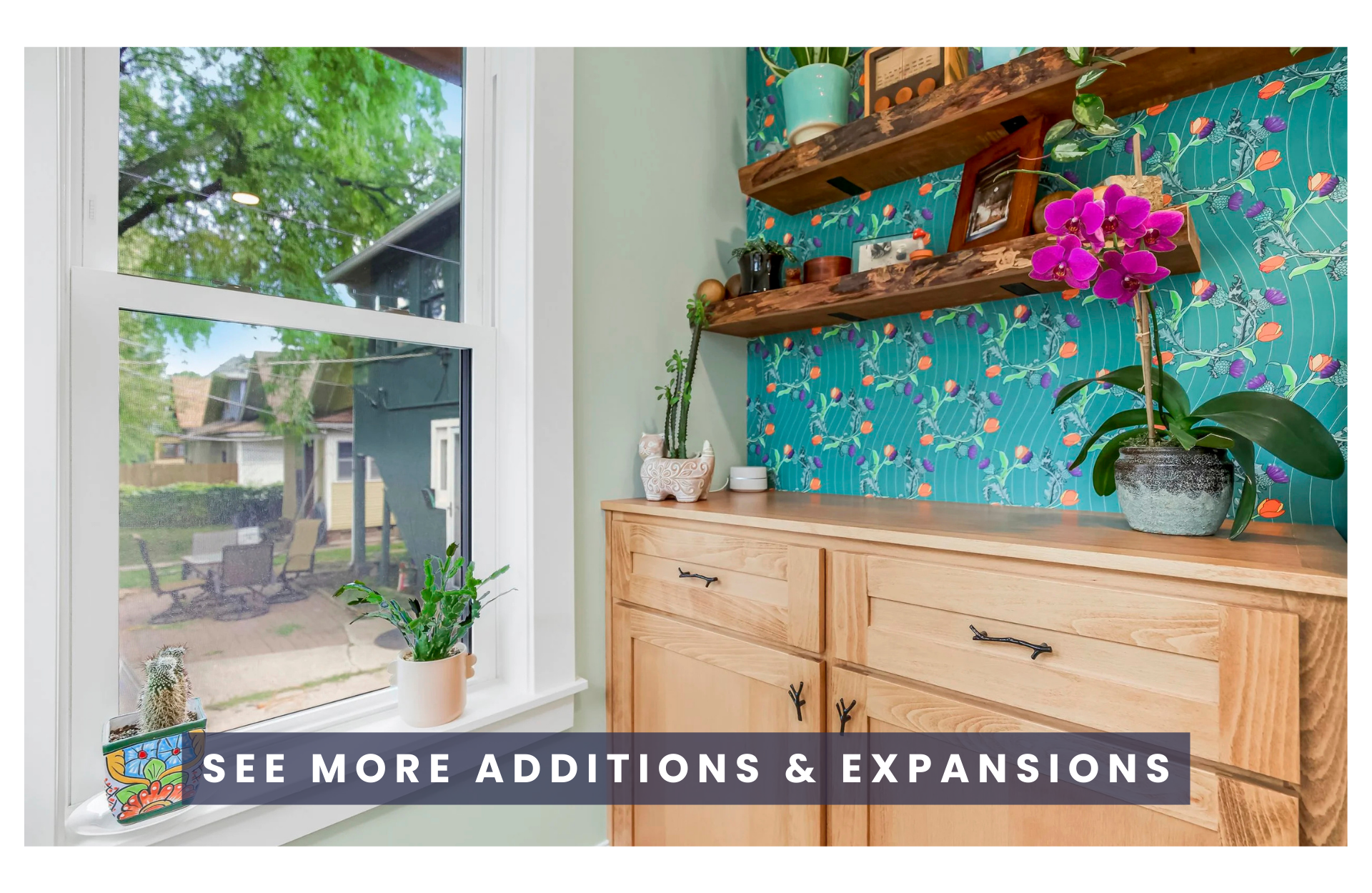 Interior corner of a laundry room with a wooden cabinet, decorative plants, and open wooden shelves against a colorful floral wallpapered wall, near a large window showing an outdoor patio with trees and outdoor furniture.