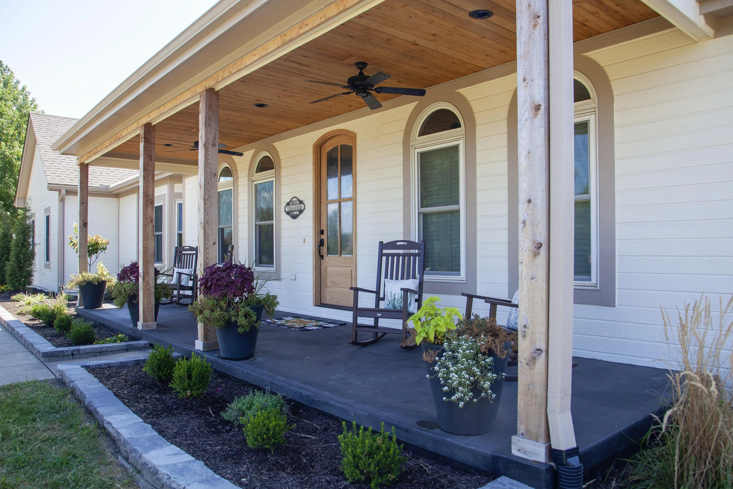 Front porch of a house with potted plants, rocking chairs, and a wooden door, featuring a wooden ceiling with ceiling fans.